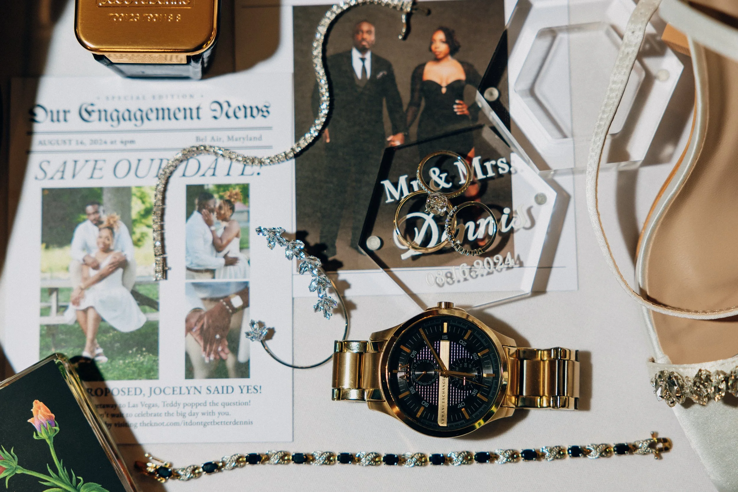 Wedding memorabilia on a table, including a Rolex watch, jewelry, rings in a display box, a wedding announcement, a smartphone, and photographs of a couple.