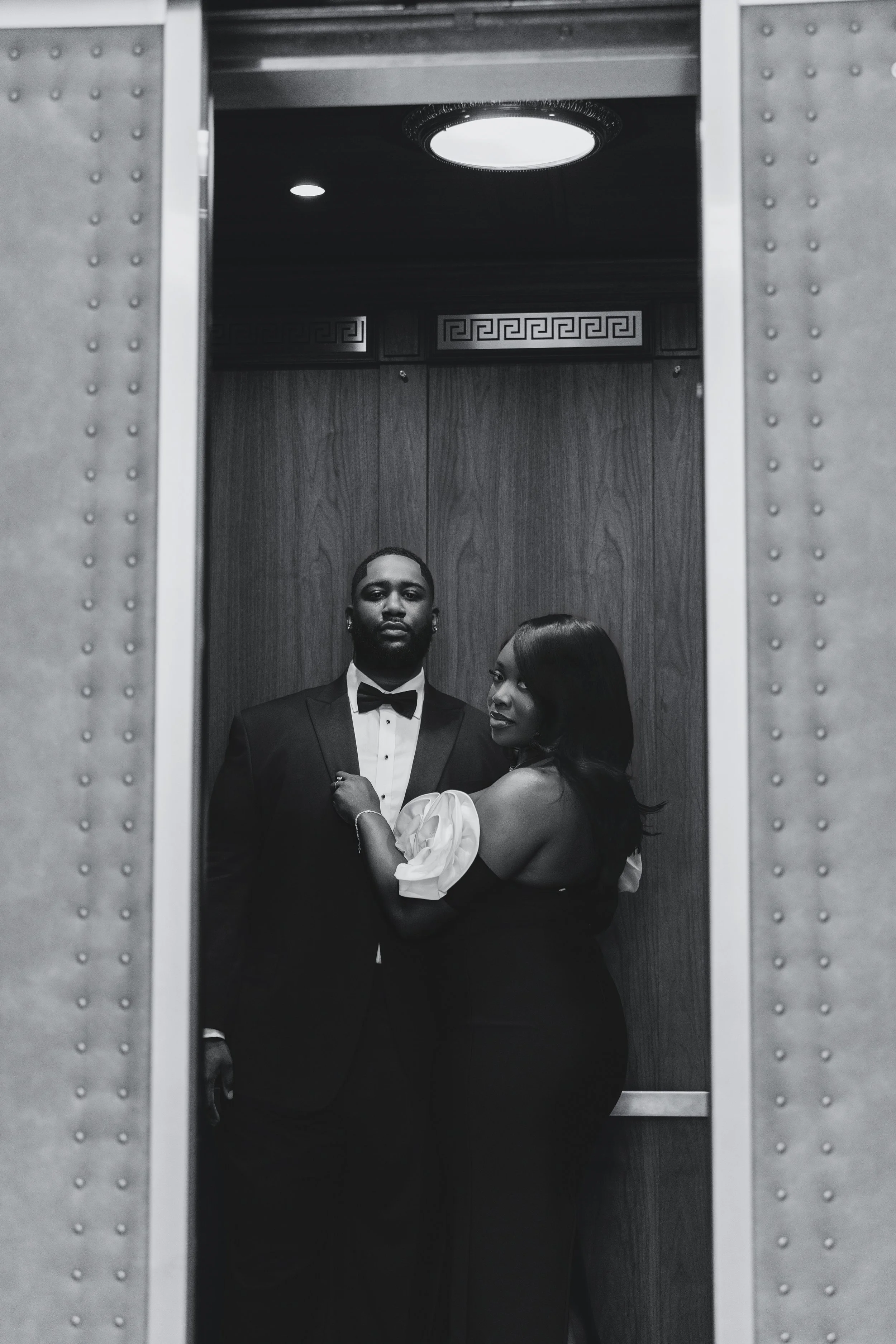 A black-and-white photo of a man in a tuxedo and wearing a bow tie standing beside a woman in an elegant black dress with puffed sleeves inside an elevator, with their reflection visible in the mirror.