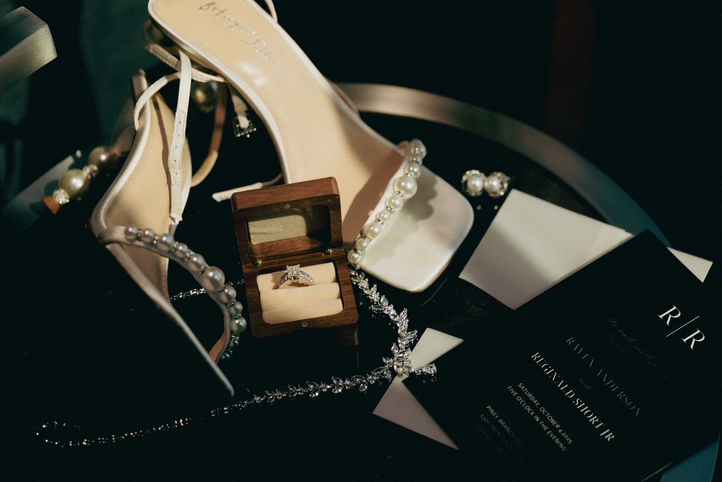 Wedding jewelry including a pearl necklace, pearl earrings, a diamond ring in a wooden box, and a pearl bracelet surrounding a wedding invitation.