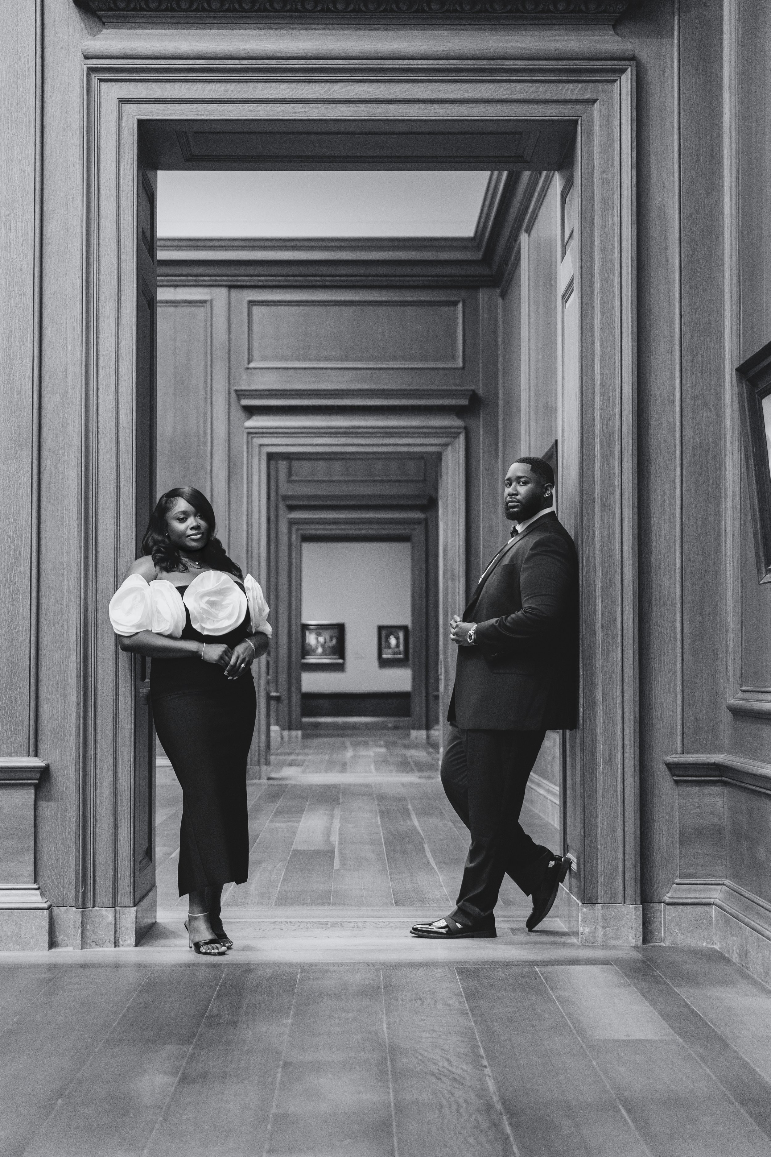 A black and white photo of a man and woman standing in a wooden-paneled hallway. The woman is on the left, dressed in an elegant dress with puffed sleeves, and the man is on the right wearing a suit. They are posing for the camera.