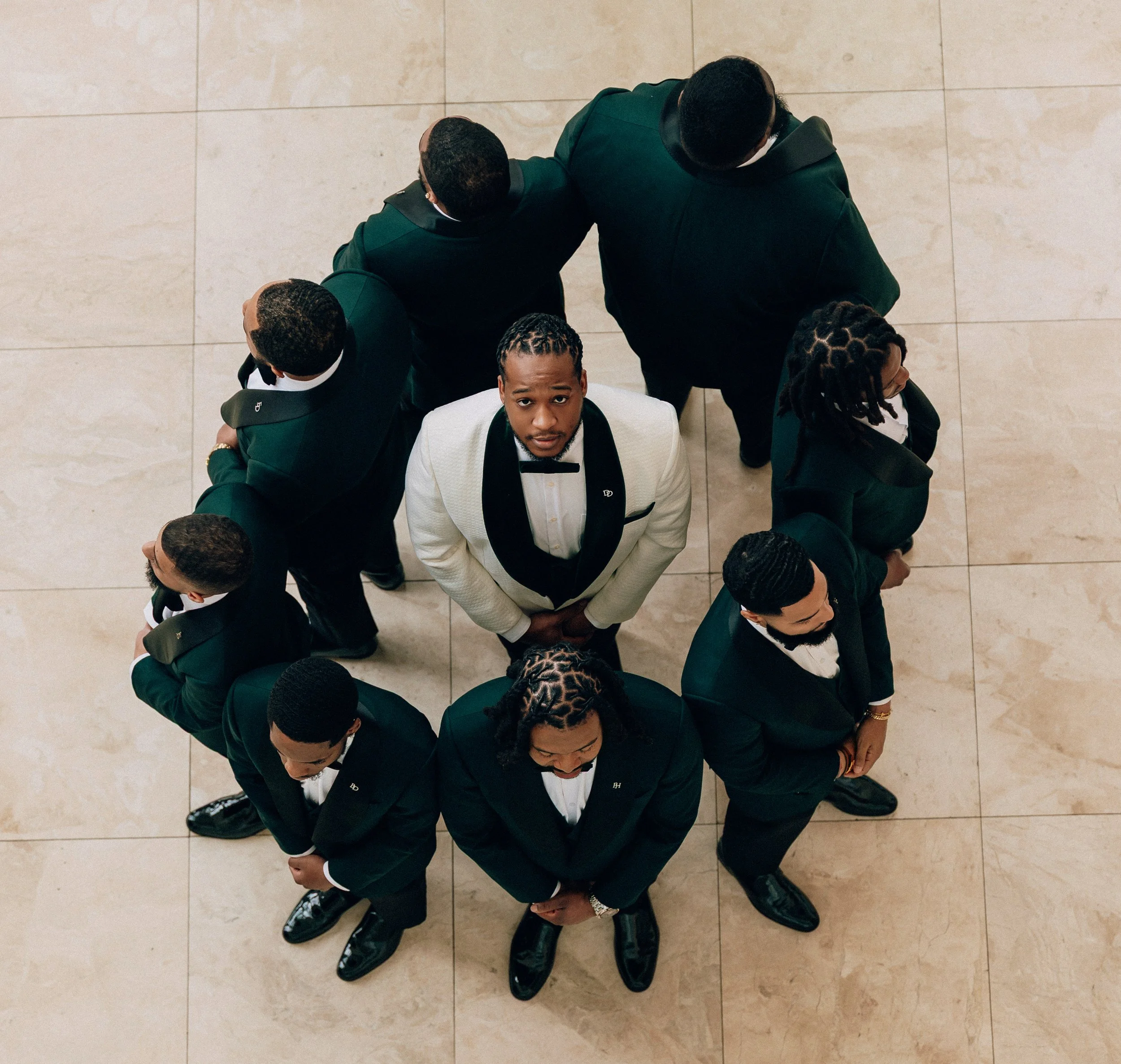 Group of men dressed in black tuxedos and a man in a white tuxedo with a black bow tie, standing on a marble tiled floor in a circle, looking up at the camera.