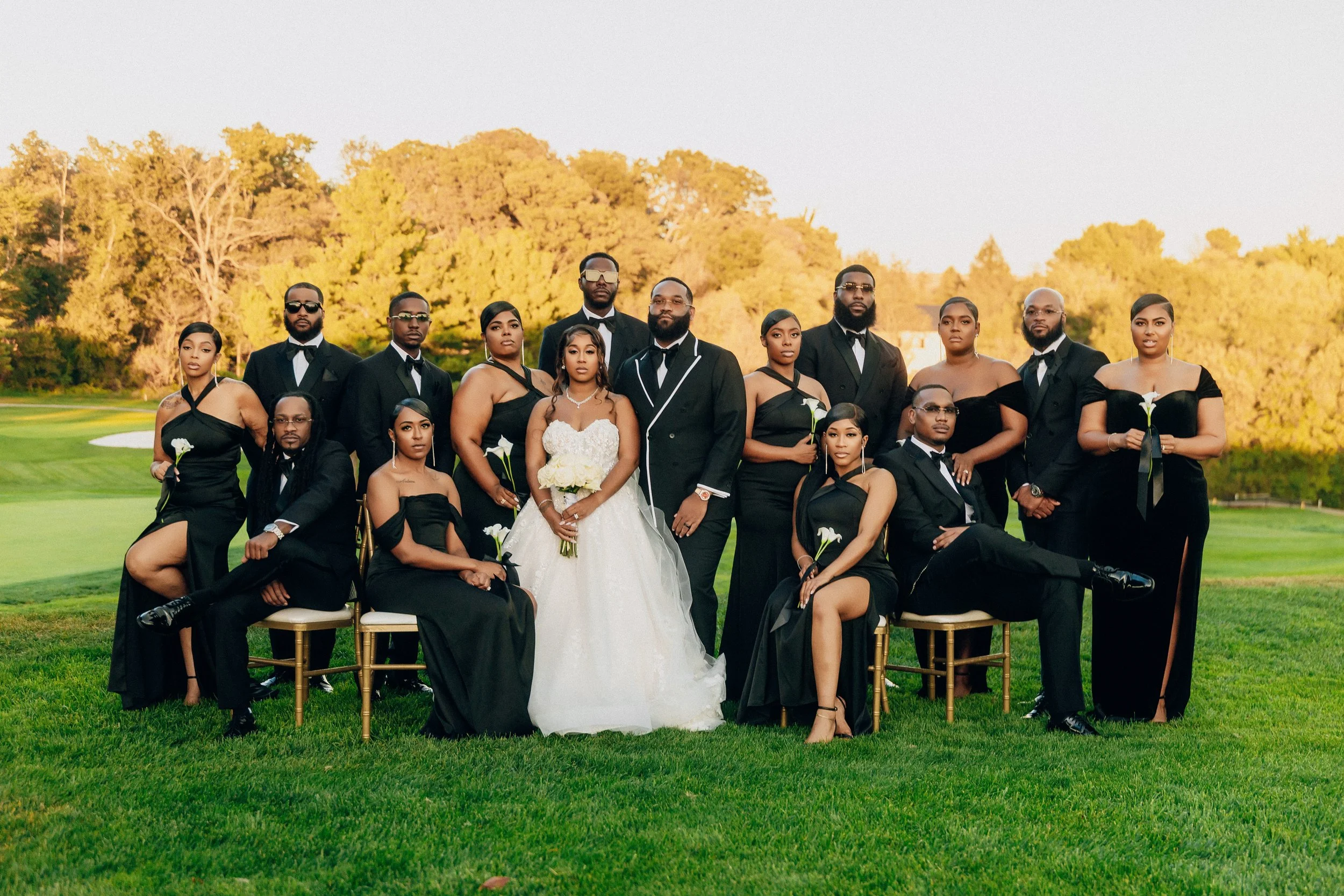 Group of people, dressed formally in black and white, posing outdoors on a golf course during sunset. The group includes men in tuxedos and women in elegant black dresses, with a bride in a white wedding gown holding a bouquet in the center.