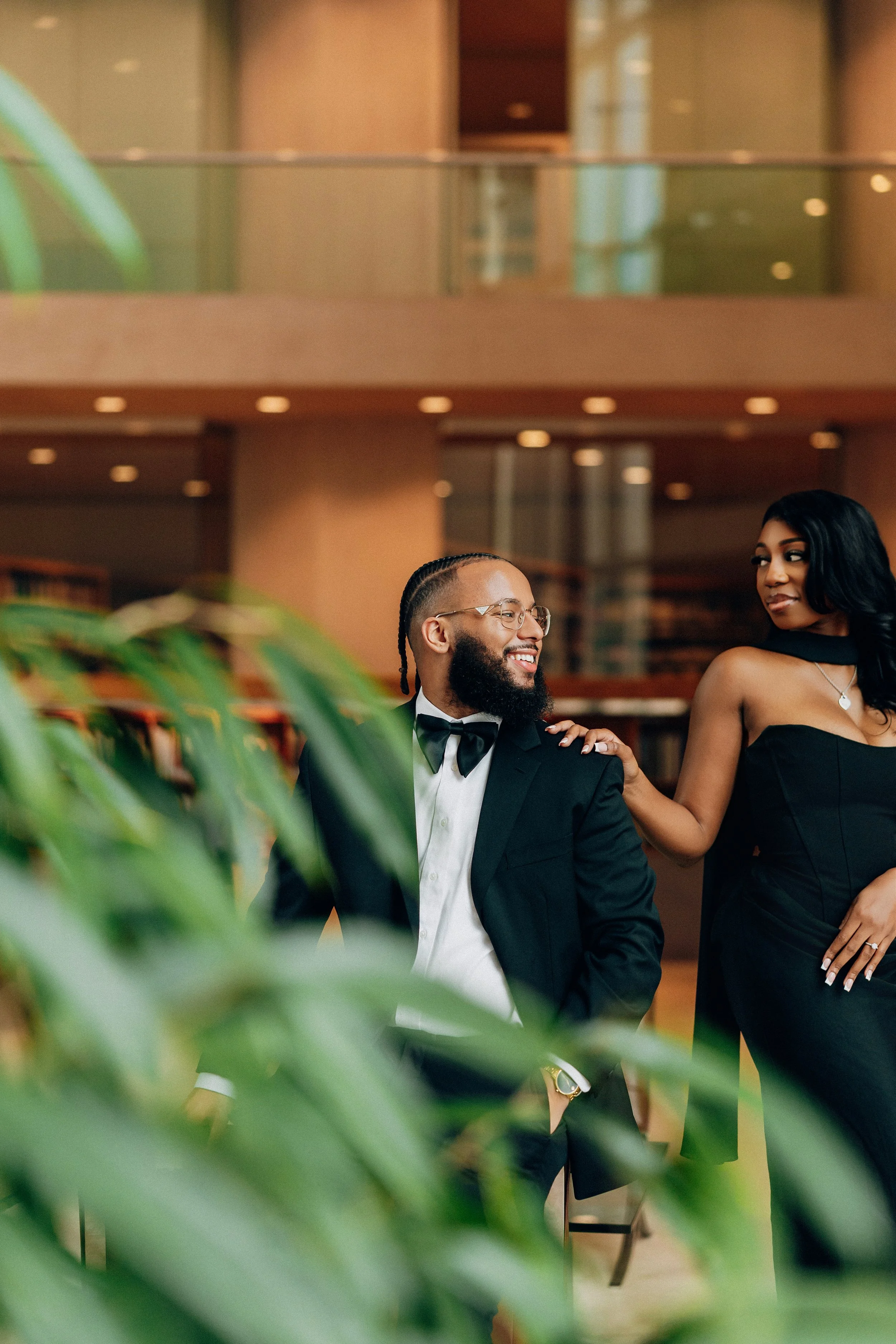 A man in a tuxedo with glasses and a woman in a black dress stand in a hotel lobby, smiling and looking at each other, with green plants partially obscuring the view.