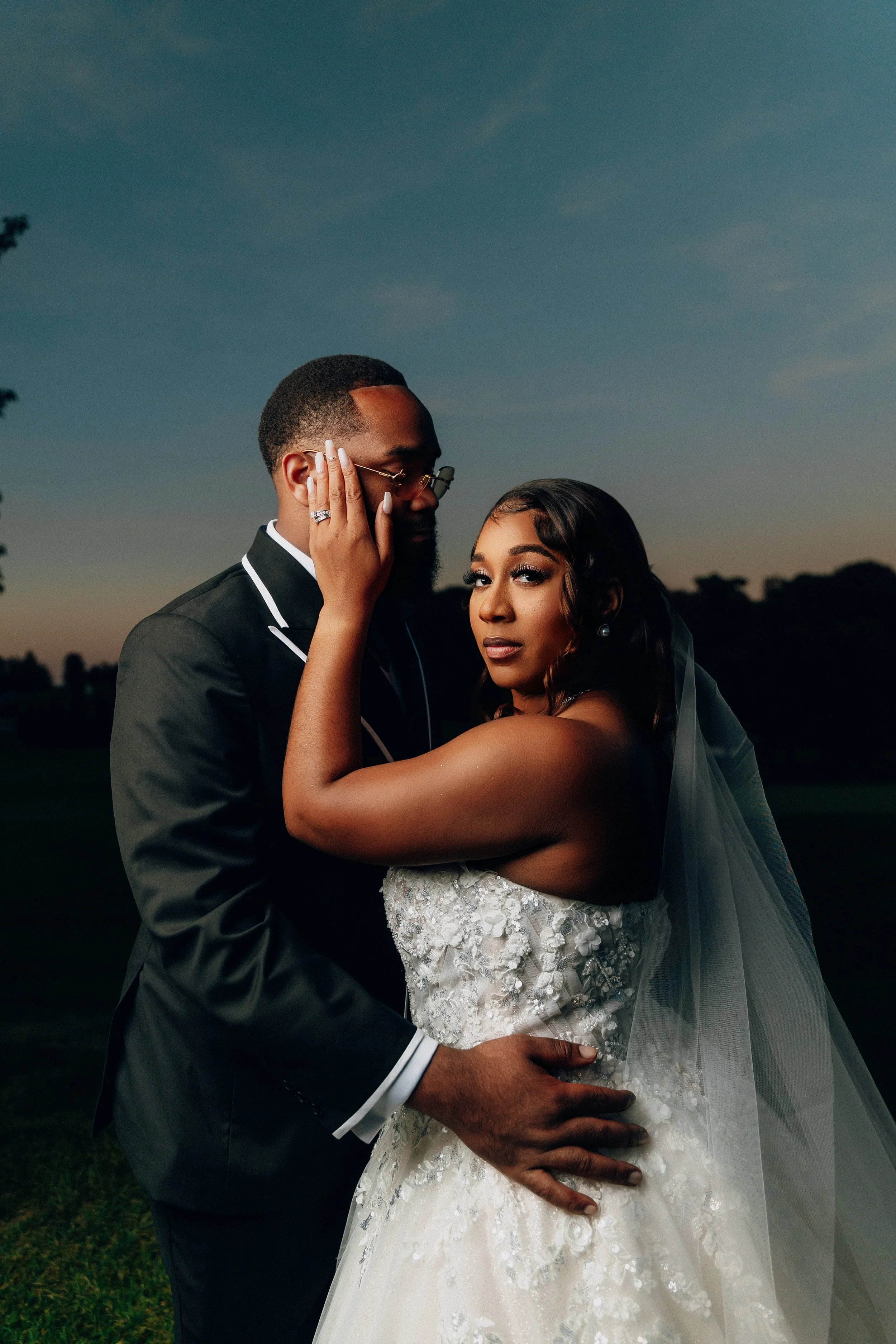 A bride and groom pose closely outdoors during sunset, with the bride wearing a white lace wedding dress and veil, and the groom in a black tuxedo, embracing each other romantically.