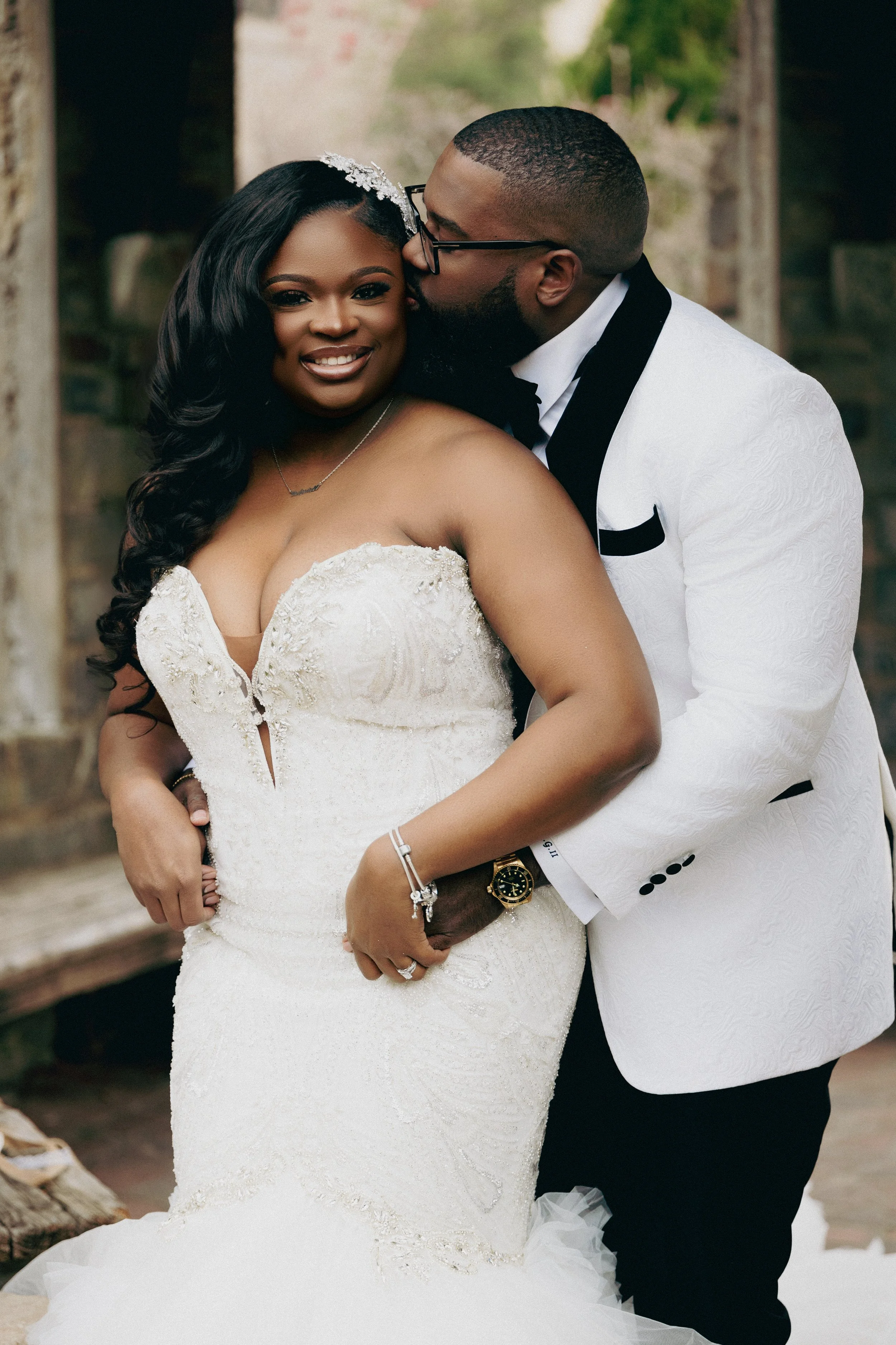 A newlywed couple dressed in wedding attire, the bride in a strapless white gown with lace details and the groom in a white tuxedo with black accents, sharing an intimate moment outdoors.