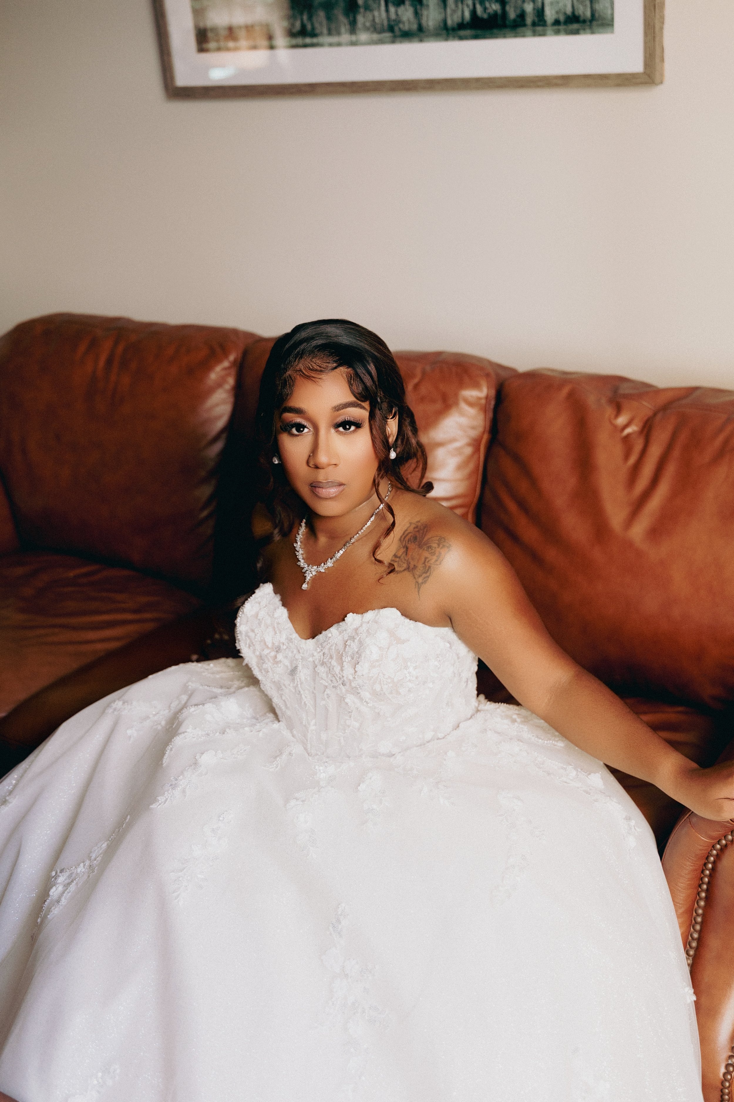 A bride sitting on a brown leather couch, wearing a white wedding dress and jewelry, looking at the camera.