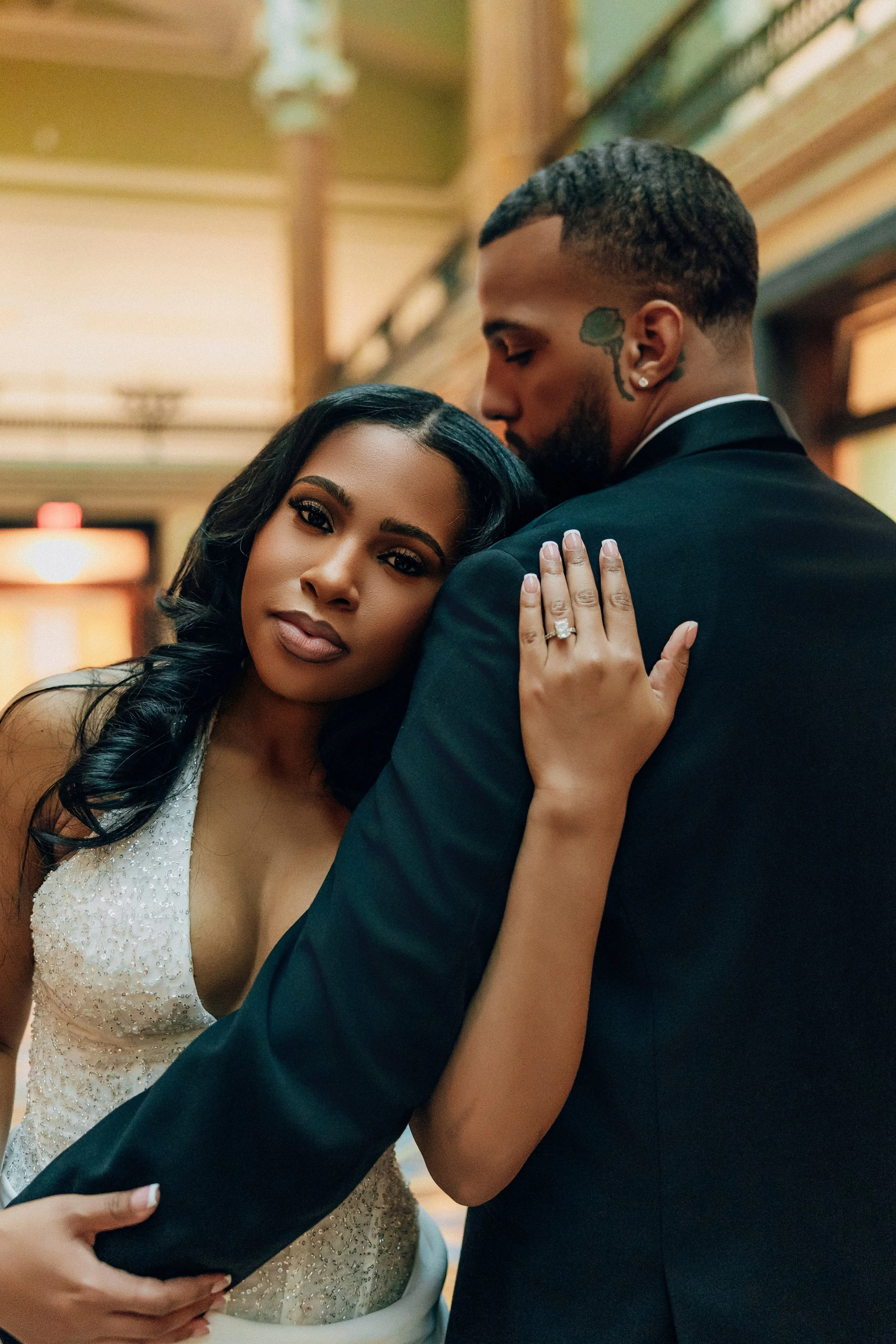 A loving couple embraces at their wedding, the woman resting her head on the man's shoulder, both dressed elegantly, in an indoor setting with warm lighting.