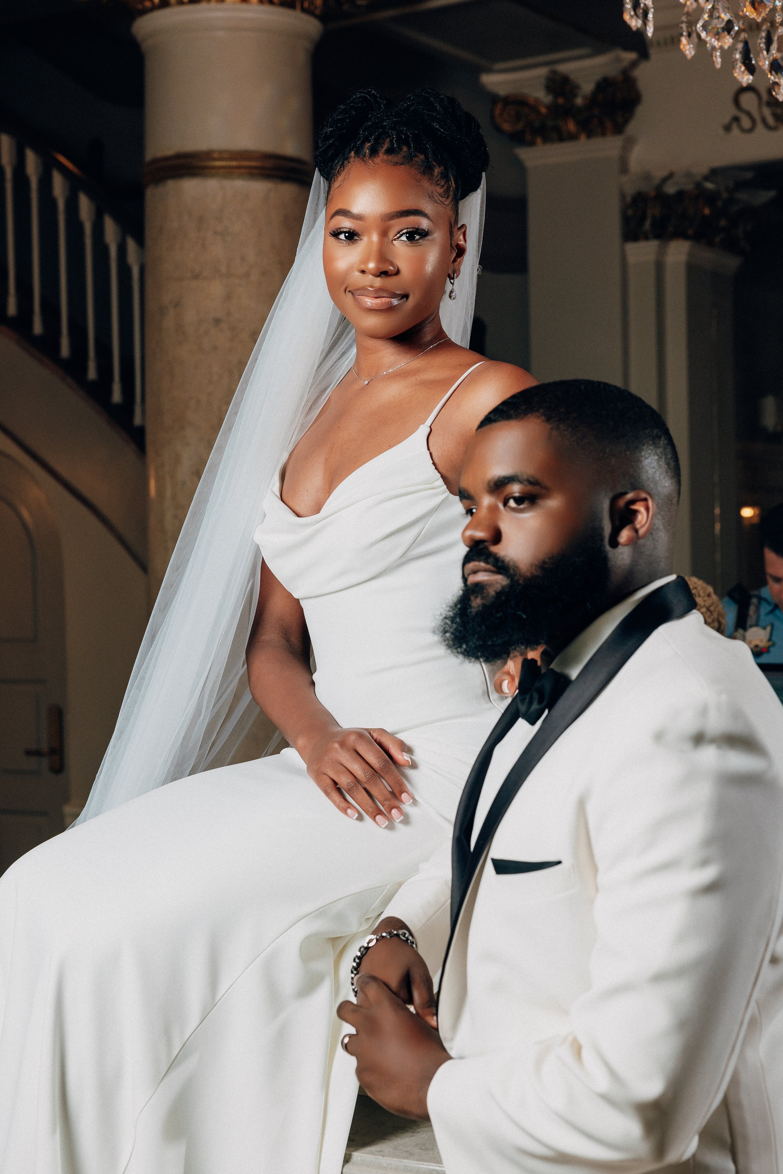 A bridal couple at their wedding, the bride wearing a white dress with a veil and the groom in a white tuxedo with a black bow tie, in an ornate indoor setting.