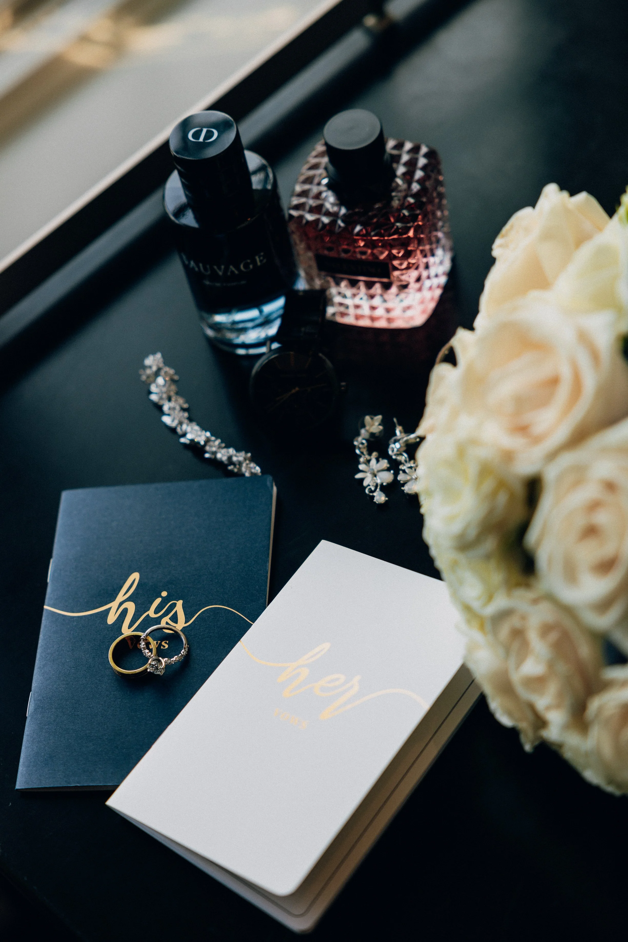 Wedding rings on a navy blue and white cards with the word 'hey' written in gold, beside a bouquet of white roses and jewelry, on a black table with perfume bottles.