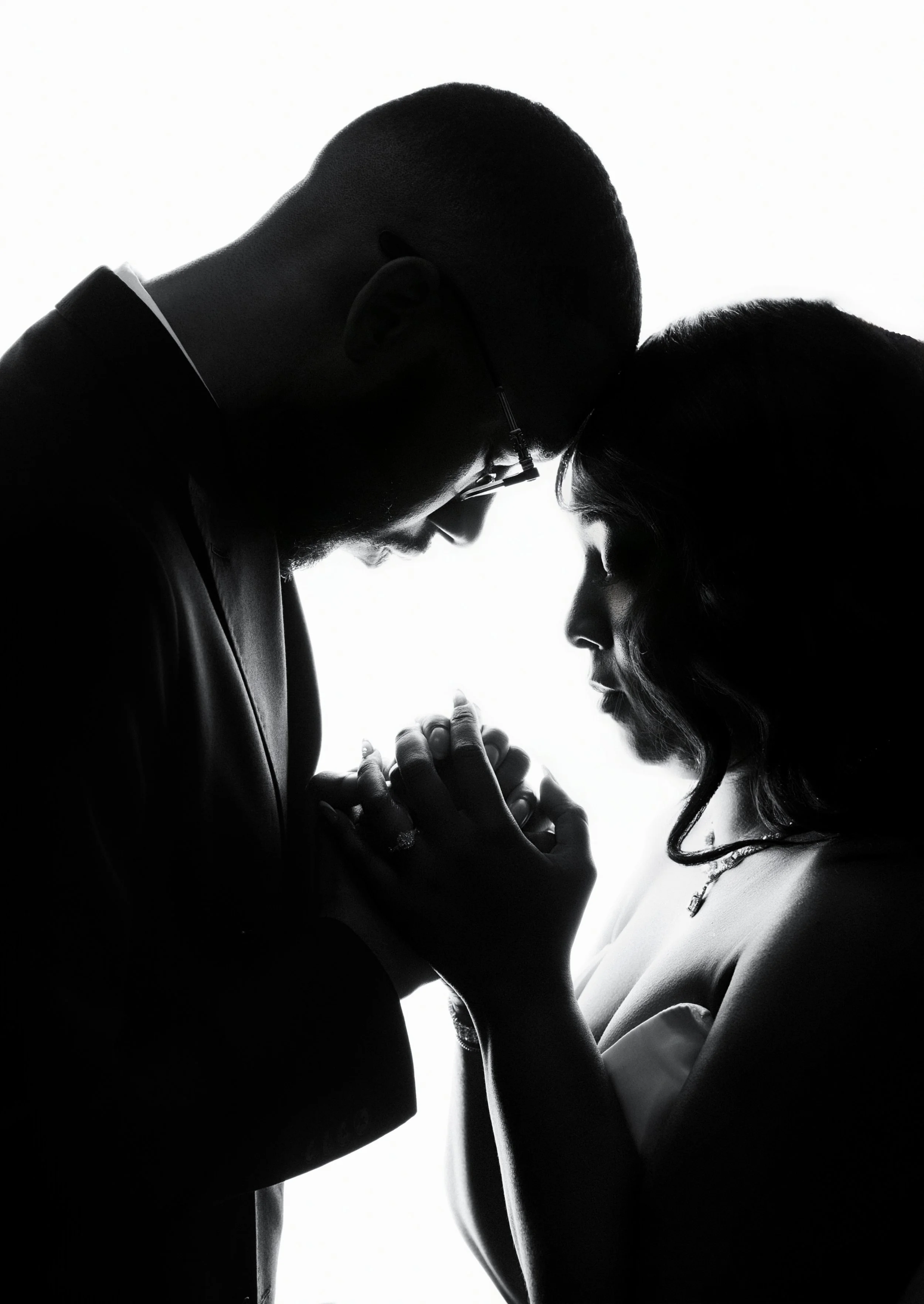 Silhouette of a couple touching foreheads and holding hands, with backlit studio lighting creating a high-contrast black and white portrait.