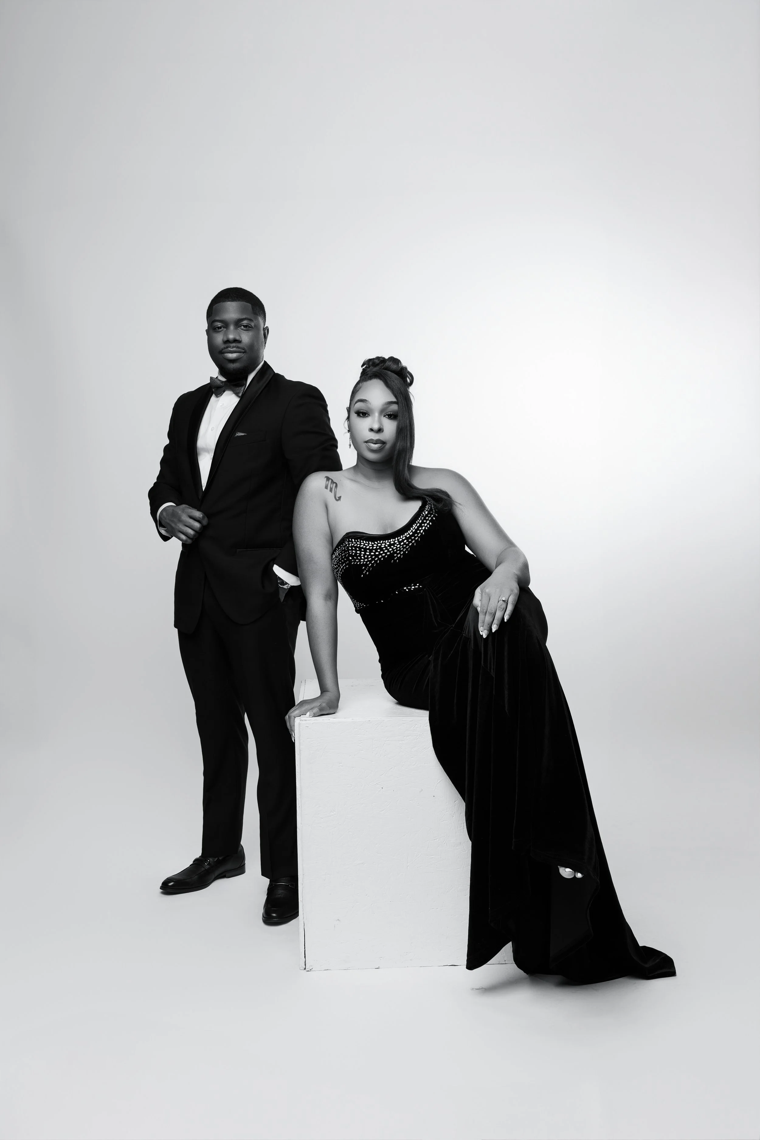 A black and white portrait of a stylishly dressed couple, with the man in a tuxedo standing and the woman in a long evening gown sitting on a white block, against a plain light background.