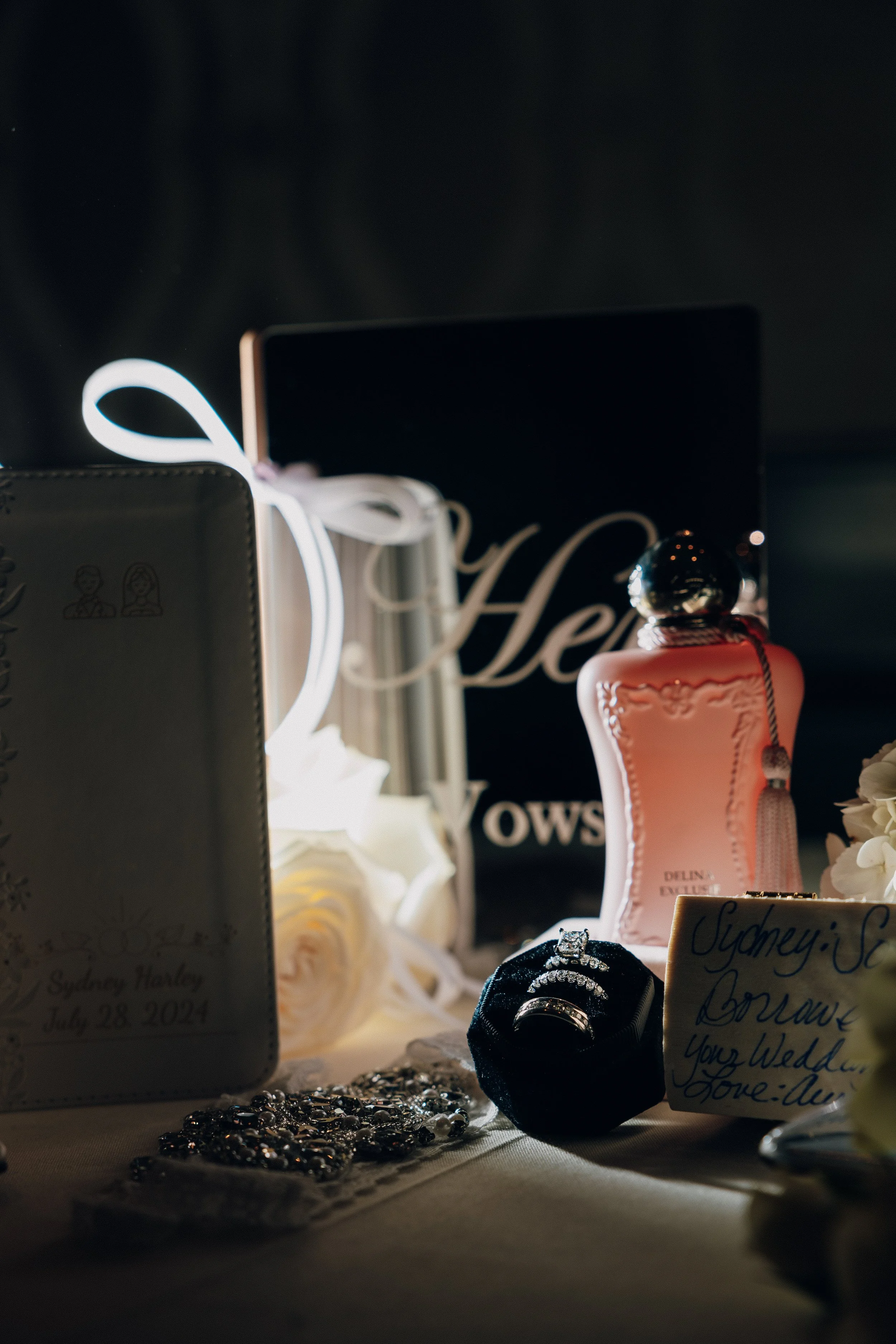 A close-up of wedding memorabilia including jewelry, a perfume bottle, and a note, with a blurred background featuring a 'Hello' sign.