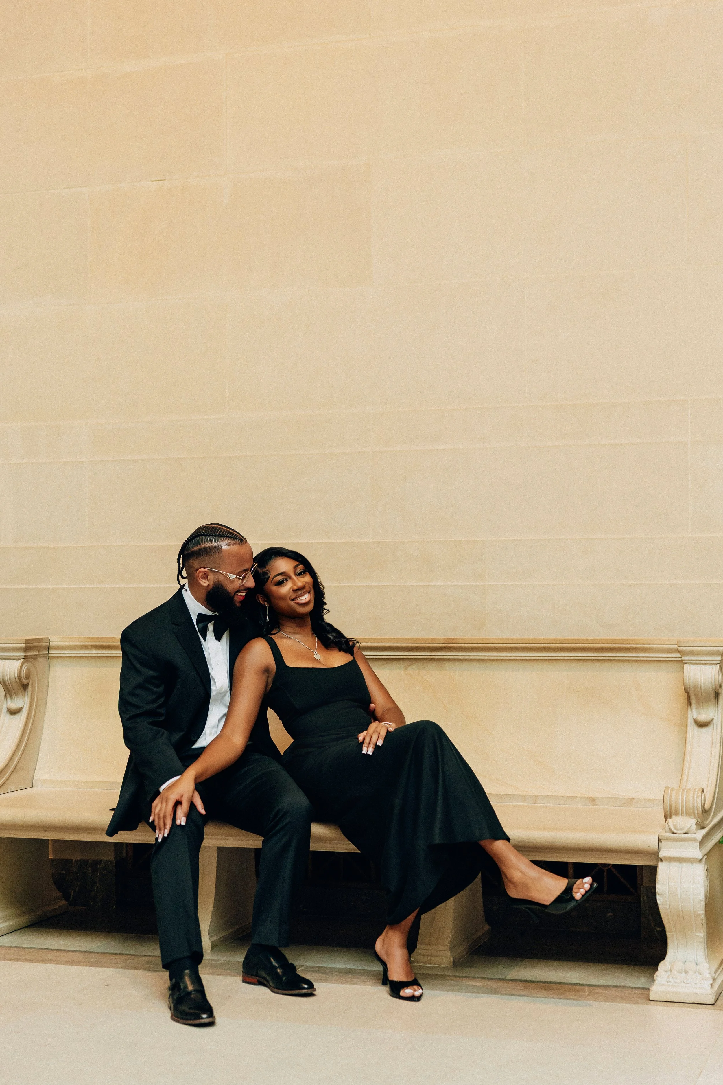 A man and woman in formal attire sitting on a wooden bench, smiling and laughing together.