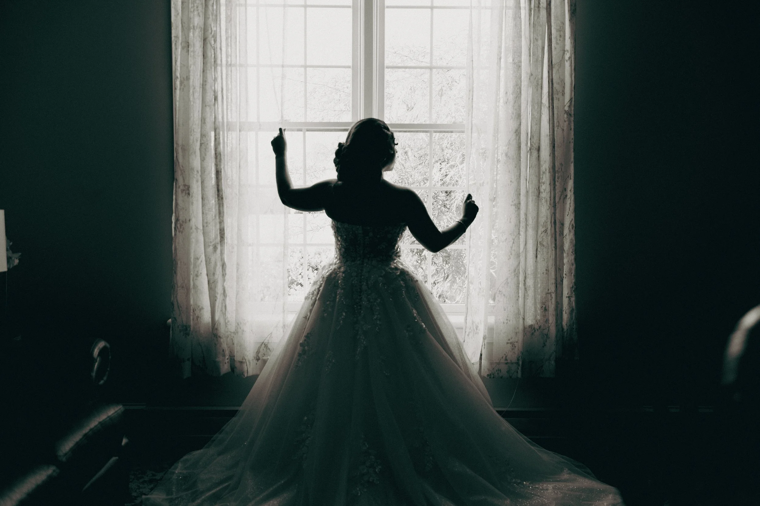 Silhouette of a woman in a wedding dress standing in front of a large window with curtains, with sunlight shining through.