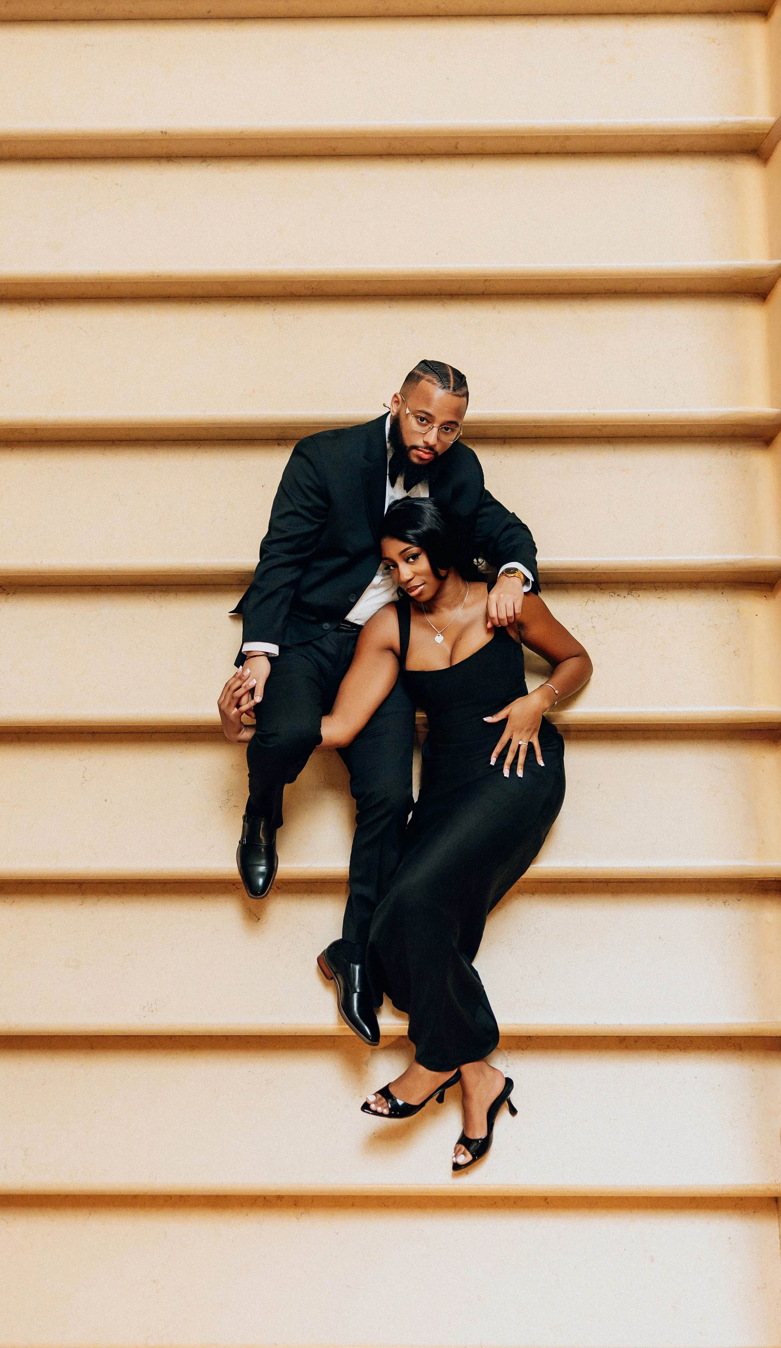 A stylish man in a black suit and blonde woman in a black dress sitting on stairs, posing together.