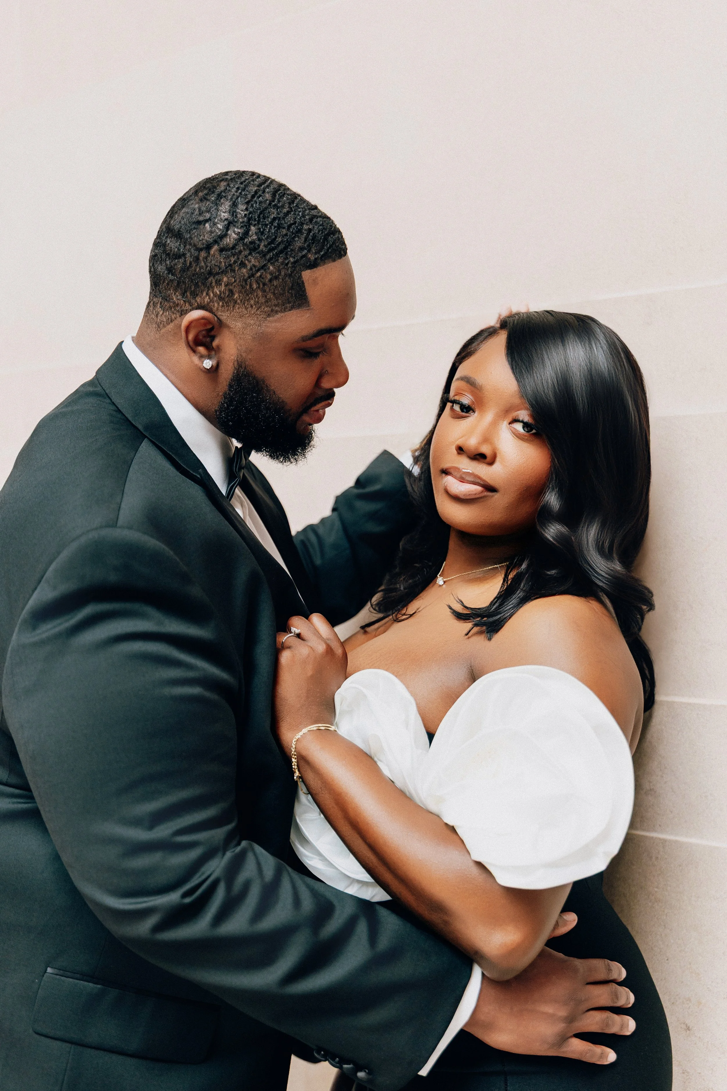 A man in a black tuxedo with a black bow tie and a woman in an off-shoulder white top with puffed sleeves, standing close together against a plain wall, gazing at each other.