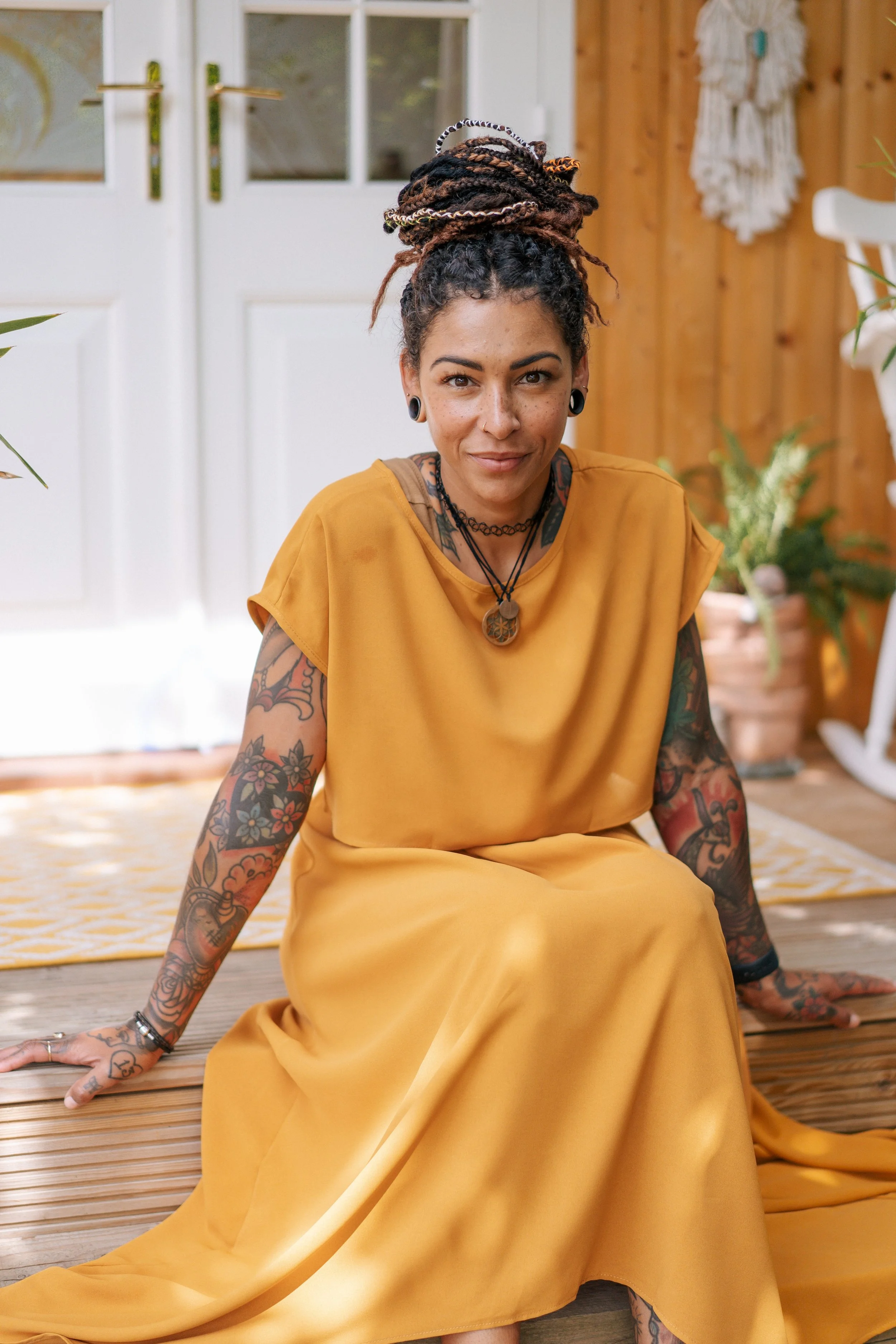 A woman with tattoos and braided hair in a bun, wearing a mustard-colored dress, sitting on a wooden floor indoors with a white door and wooden walls, surrounded by plants.
