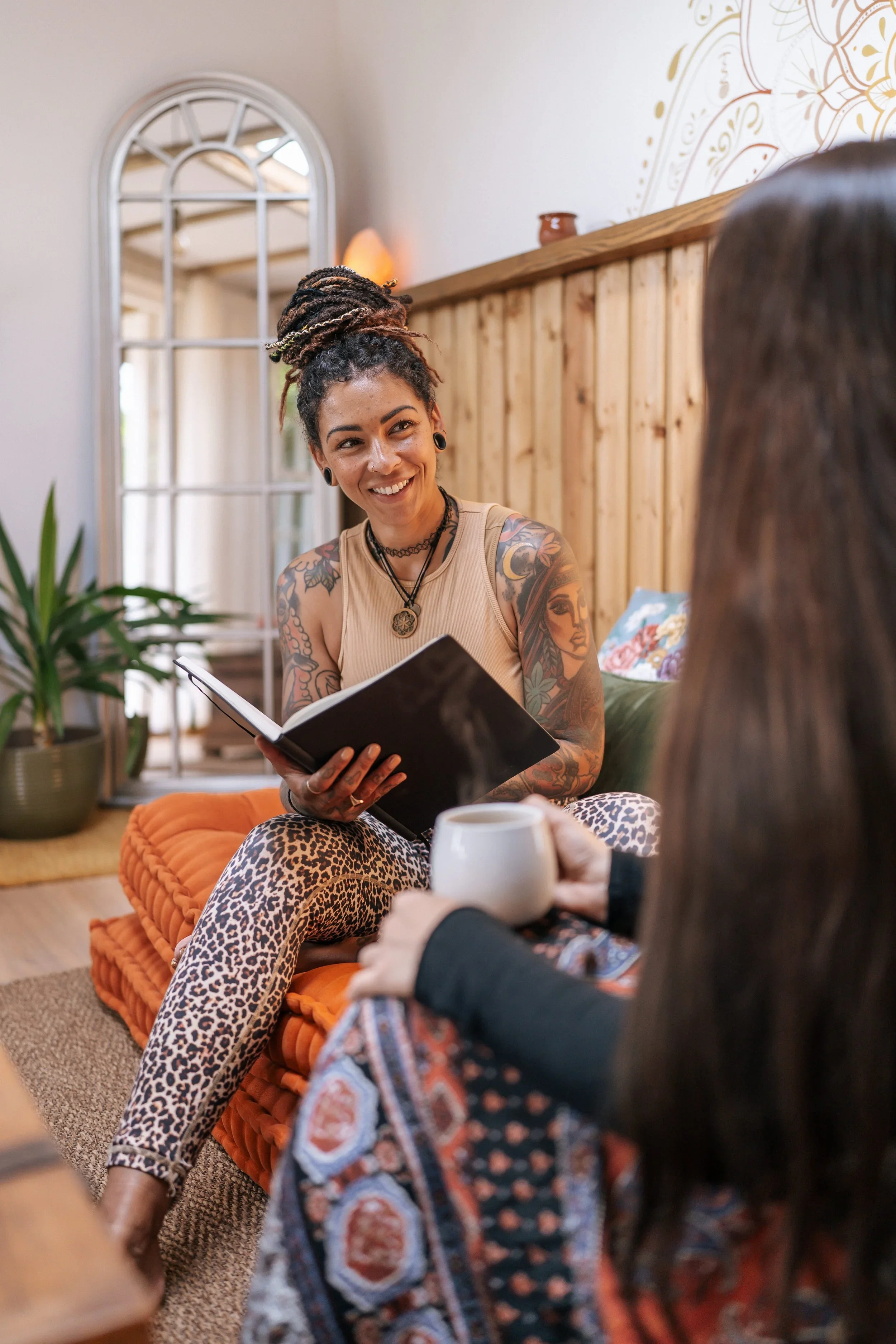 Two women are sitting in a cozy, decorated room having a conversation. One woman with tattoos, dreadlocks, and wearing a beige tank top is smiling and holding a notebook, while the other, with dark hair, is holding a white mug and facing her.