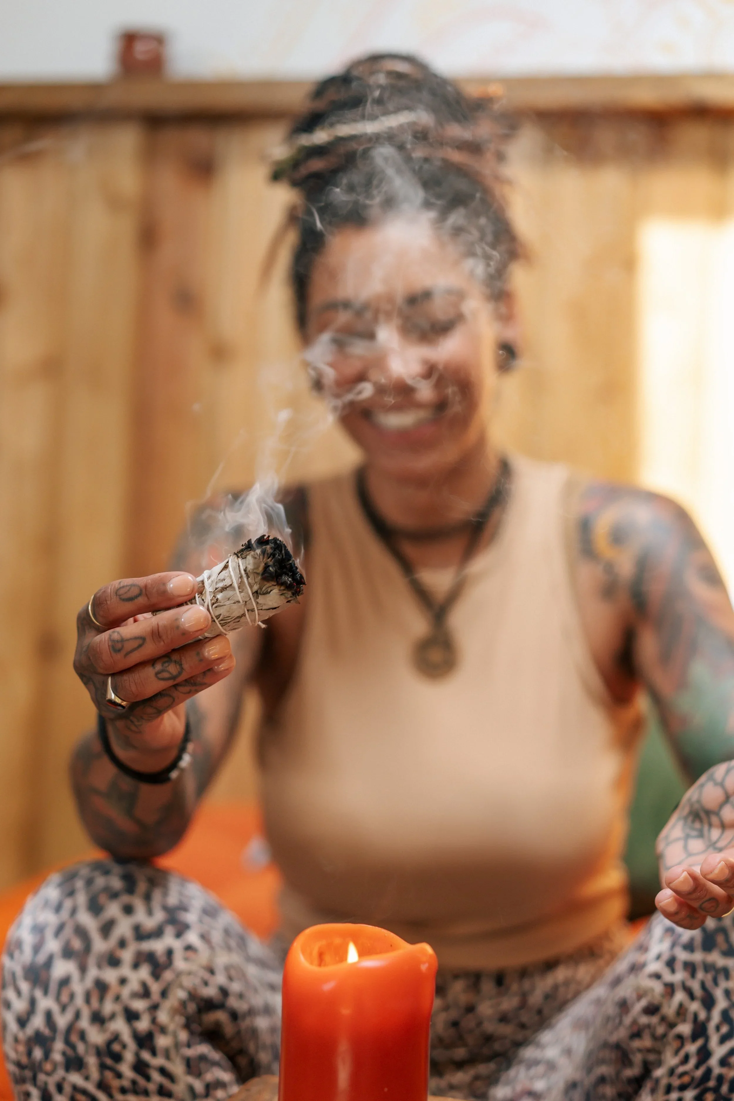 A smiling woman with tattoos on her arms, holding a burning sage bundle, with a lit candle in front of her, sitting cross-legged in a cozy wooden room.