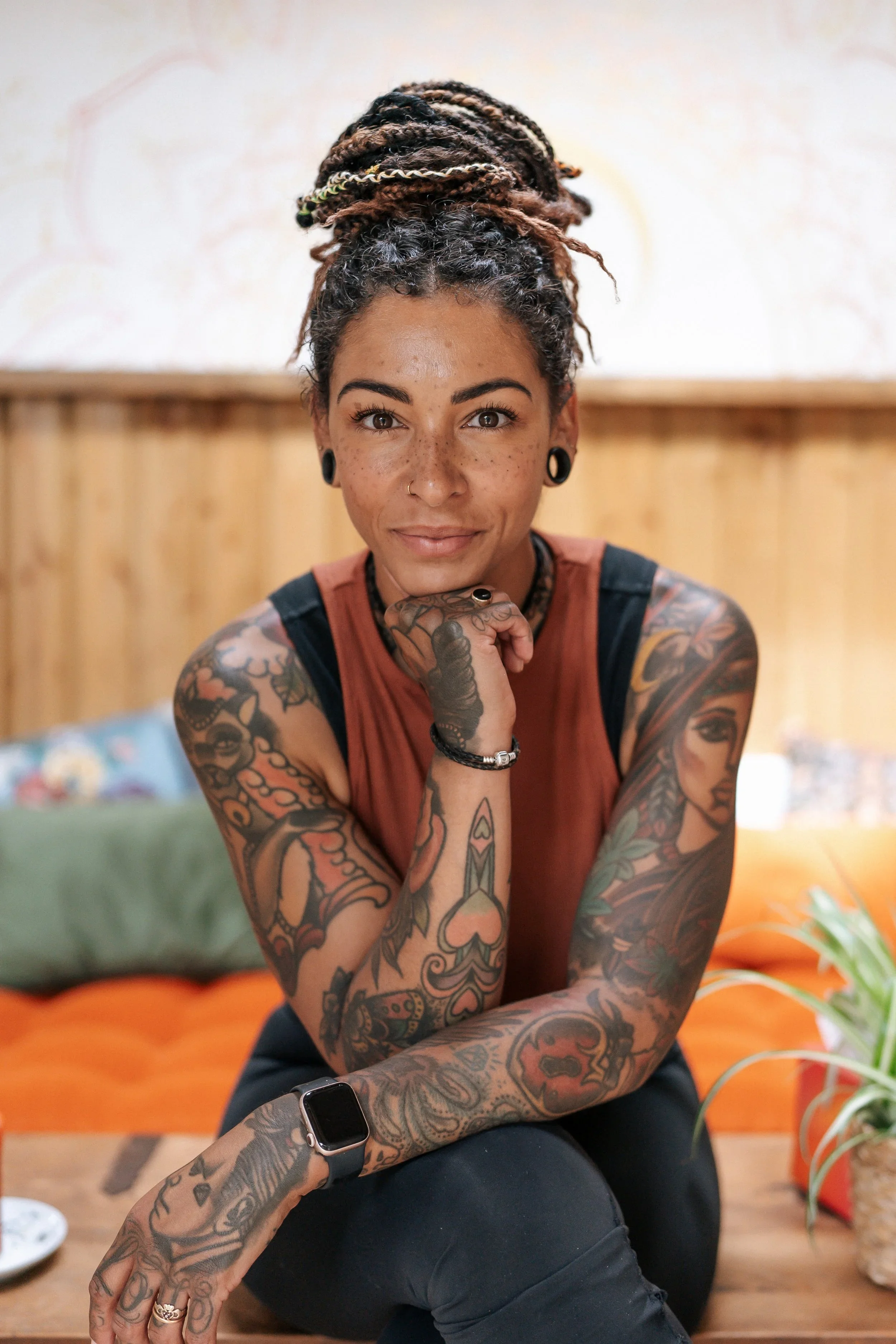 Portrait of a woman with tattoos, wearing a smartwatch, sitting indoors, with a wooden wall and plants in the background.