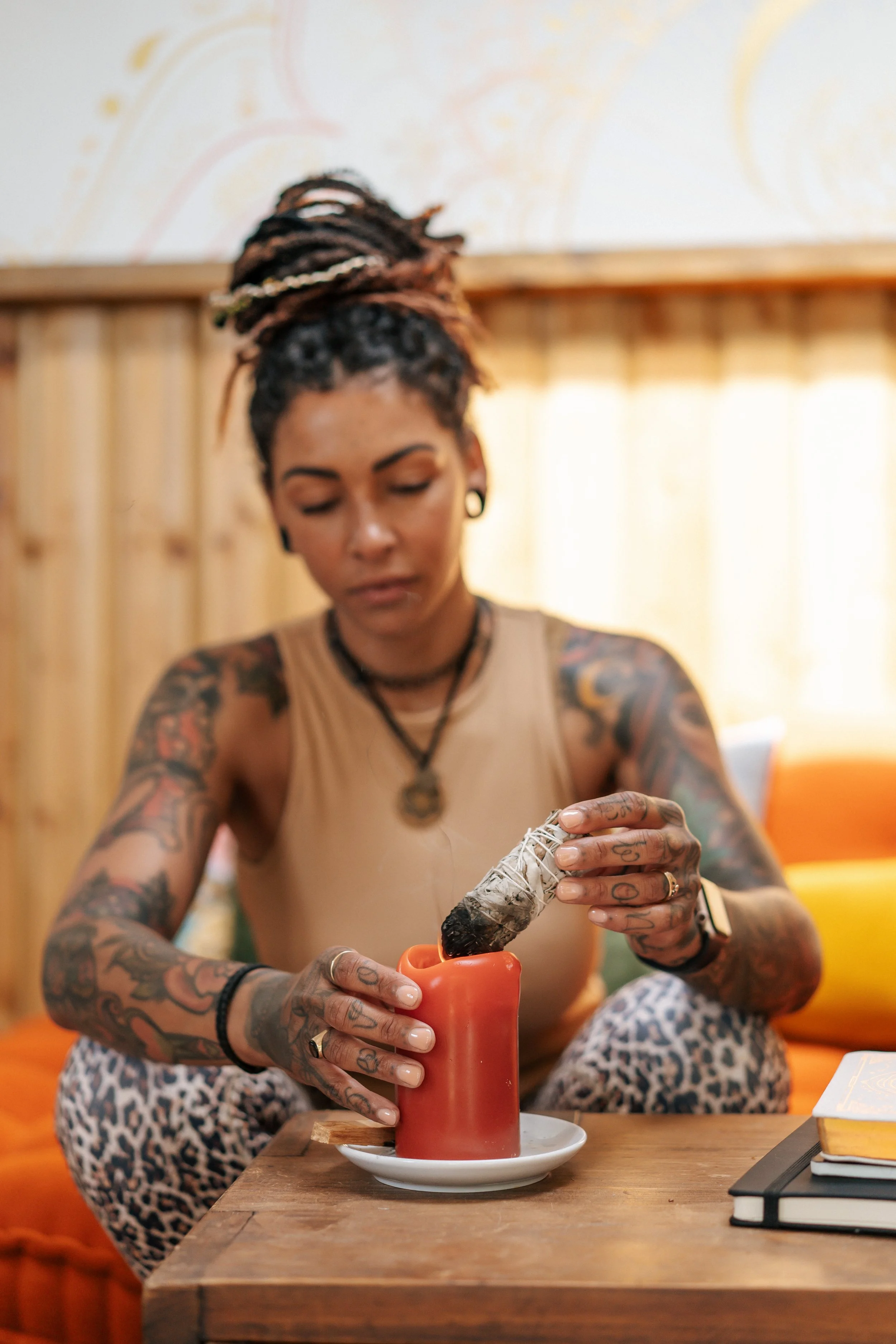 A woman with tattoos on her arms and dreadlocks is holding a burning sage bundle over a red candle and white saucer on a wooden table, in a cozy room with wooden wall paneling.