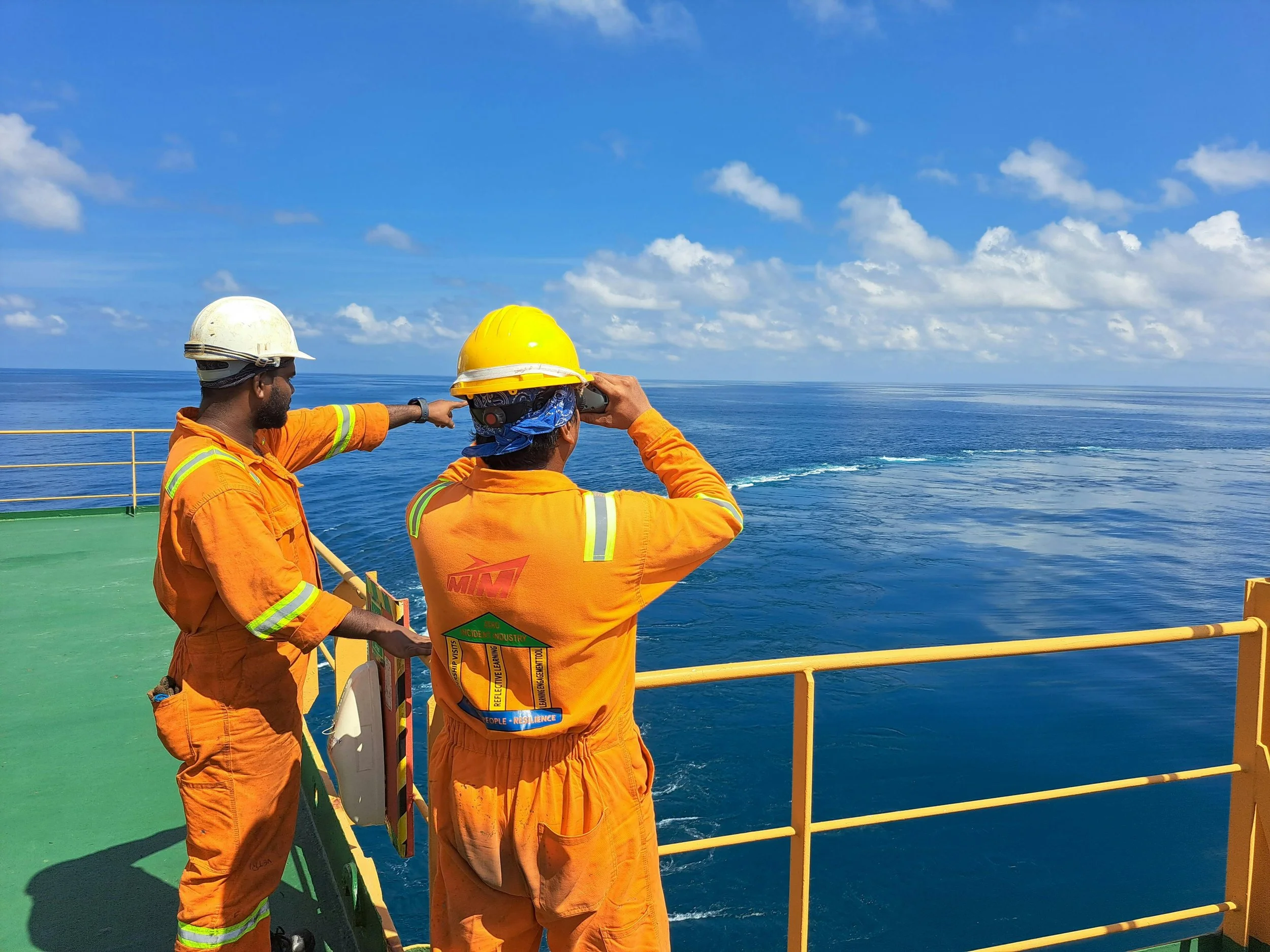 Two offshore workers in orange safety uniforms and helmets standing on a ship's deck, looking out at the ocean on a clear, sunny day.