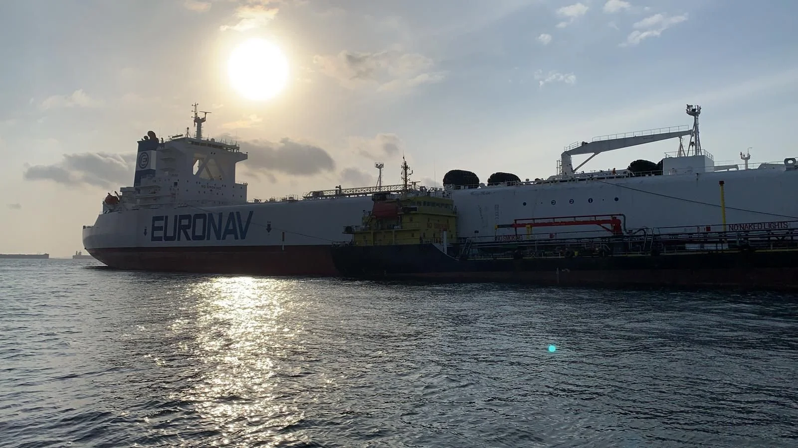 A large LNG tanker ship with the name EURONAV is docked on calm water, with the sun setting behind it and some clouds in the sky.