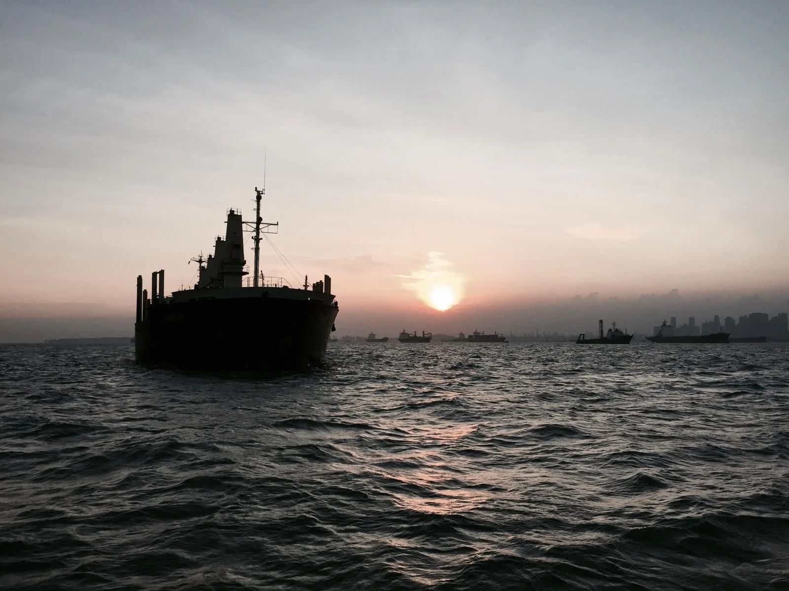 Silhouette of a large ship on the water at sunset with other ships in the background and a city skyline in the distance.