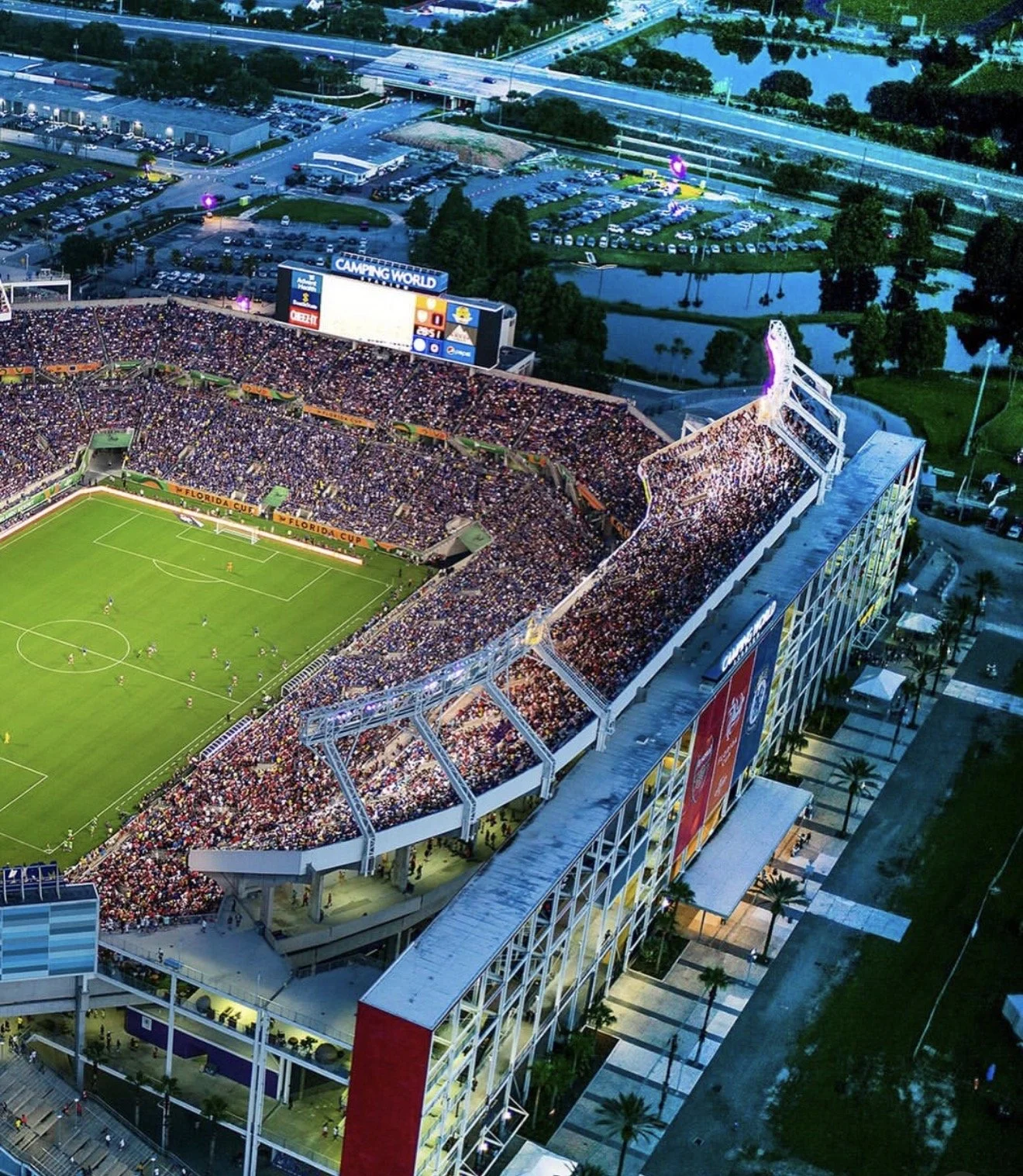 Aerial view of a packed soccer stadium during a game, with thousands of spectators in the stands and players on the field. The stadium is adjacent to parking lots, highways, and surrounding greenery, with a large digital screen displaying scores.