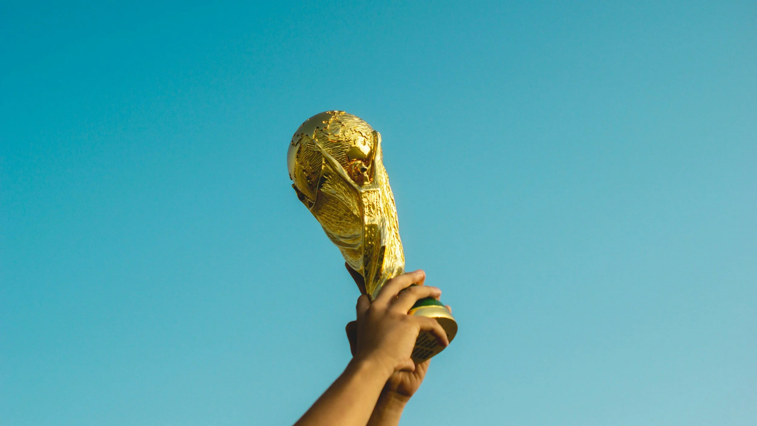 A person holding up a gold-colored FIFA World Cup trophy against a clear blue sky.