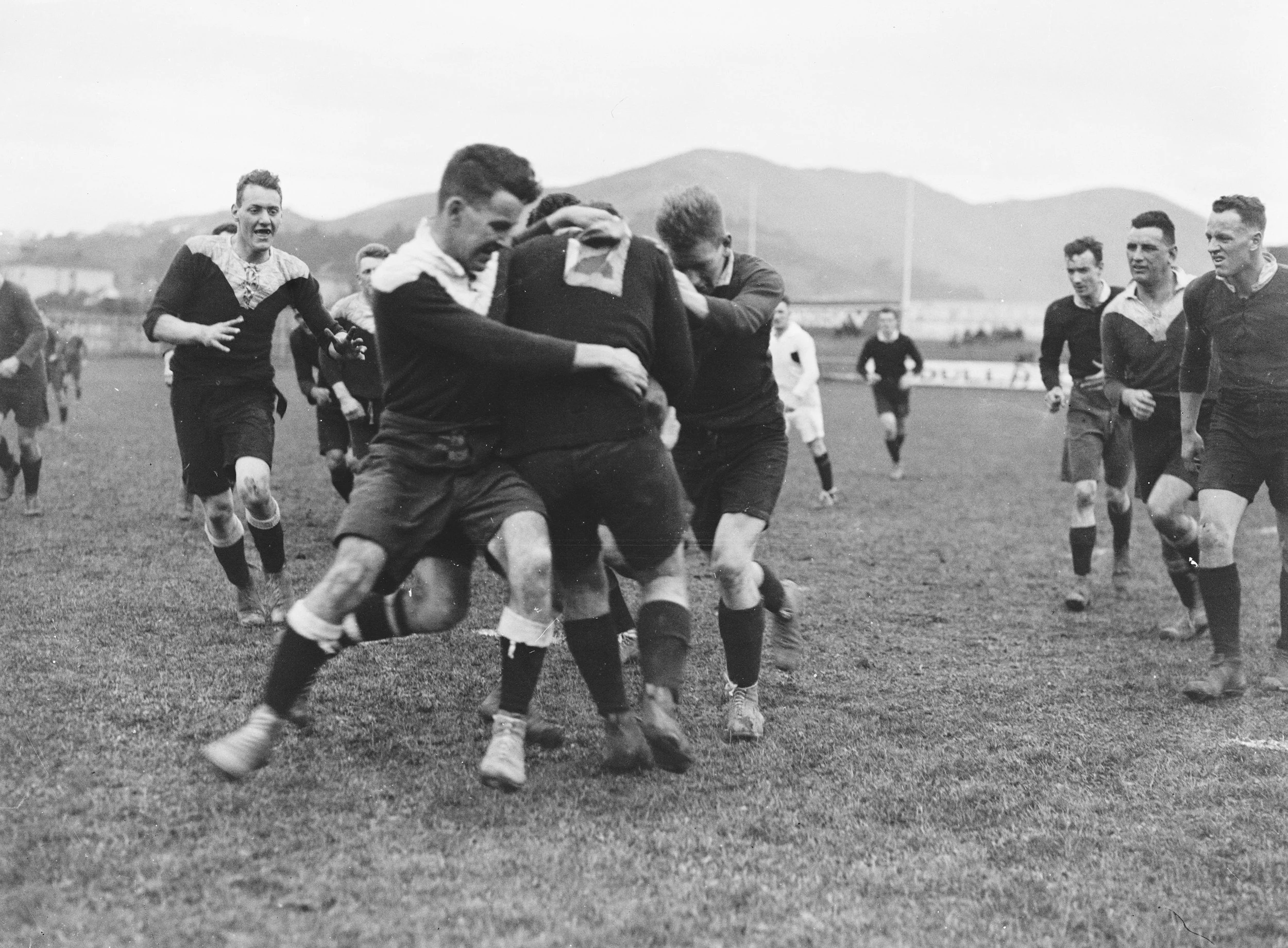 Black and white photo of a soccer game, with players running on the field, some colliding over the ball, and others trailing behind, with mountains in the background.