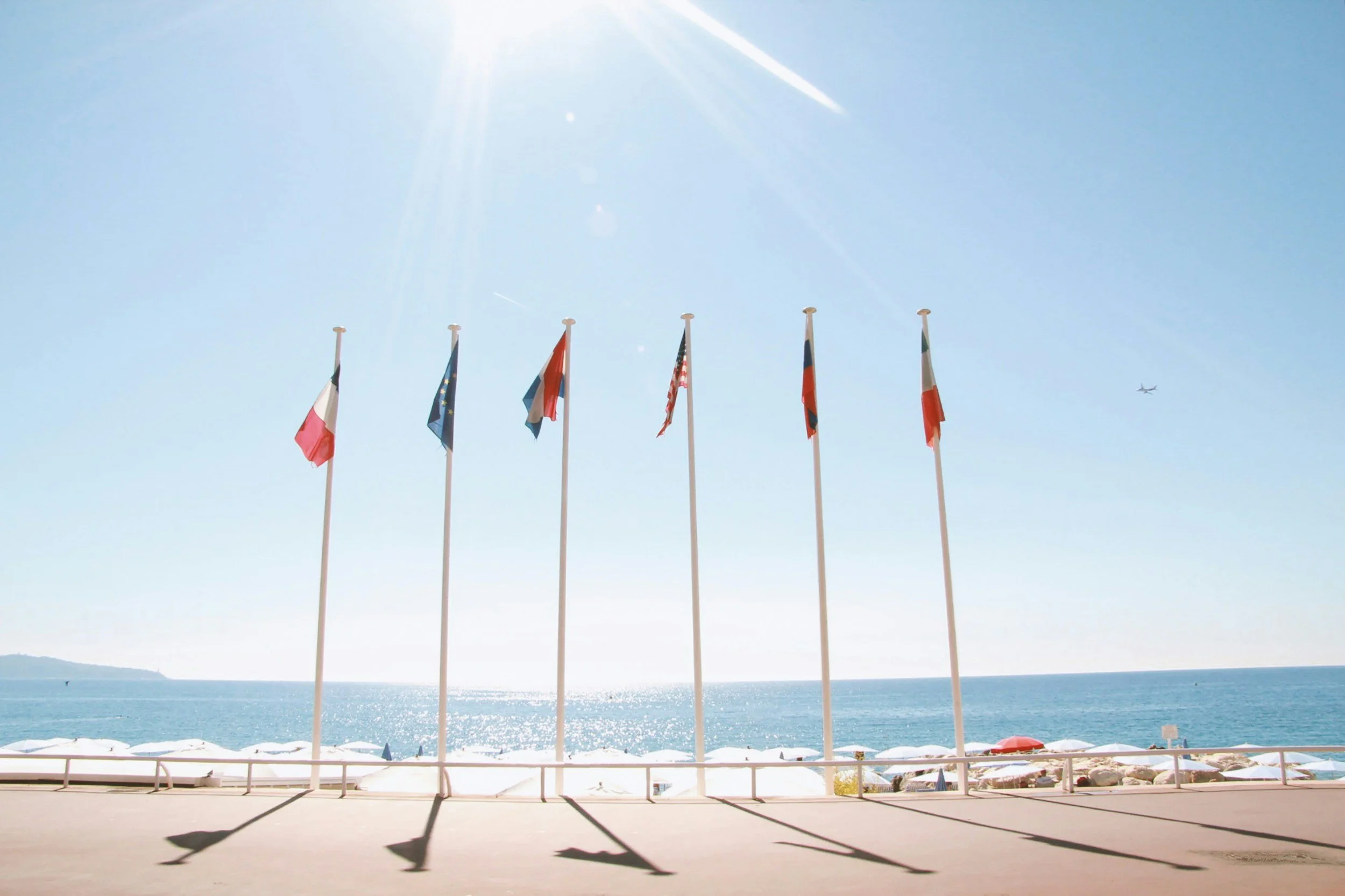 Six flags on flagpoles near a beach with umbrellas, ocean, and a clear sky with sunlight.