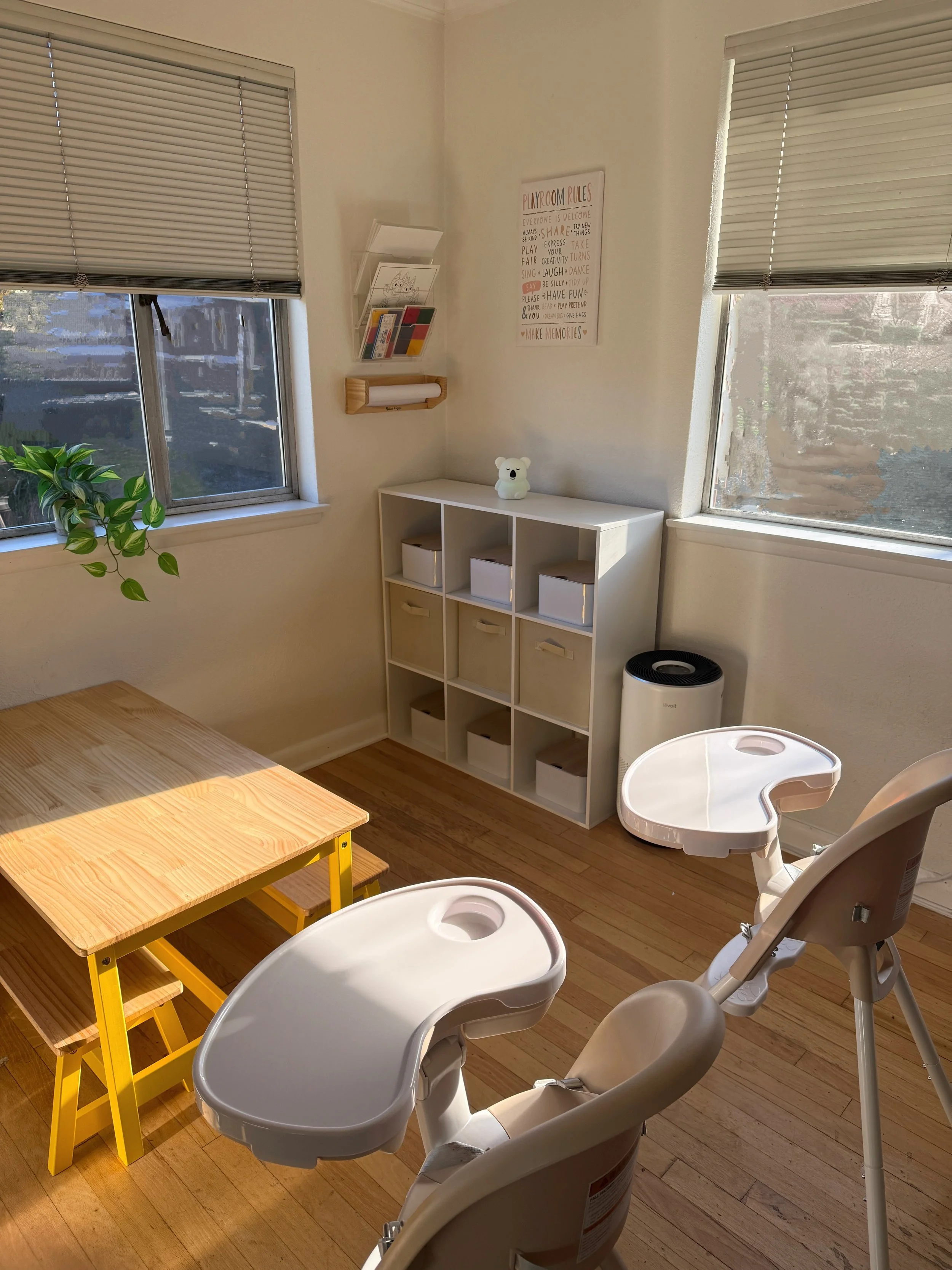 A cozy baby dining corner with two child high chairs, a small yellow table with matching stools, a white cubby storage unit with nine compartments, and a window with closed blinds. There is a small table lamp on top of the storage unit, a wall sign with playroom rules, and a white air purifier in the corner.