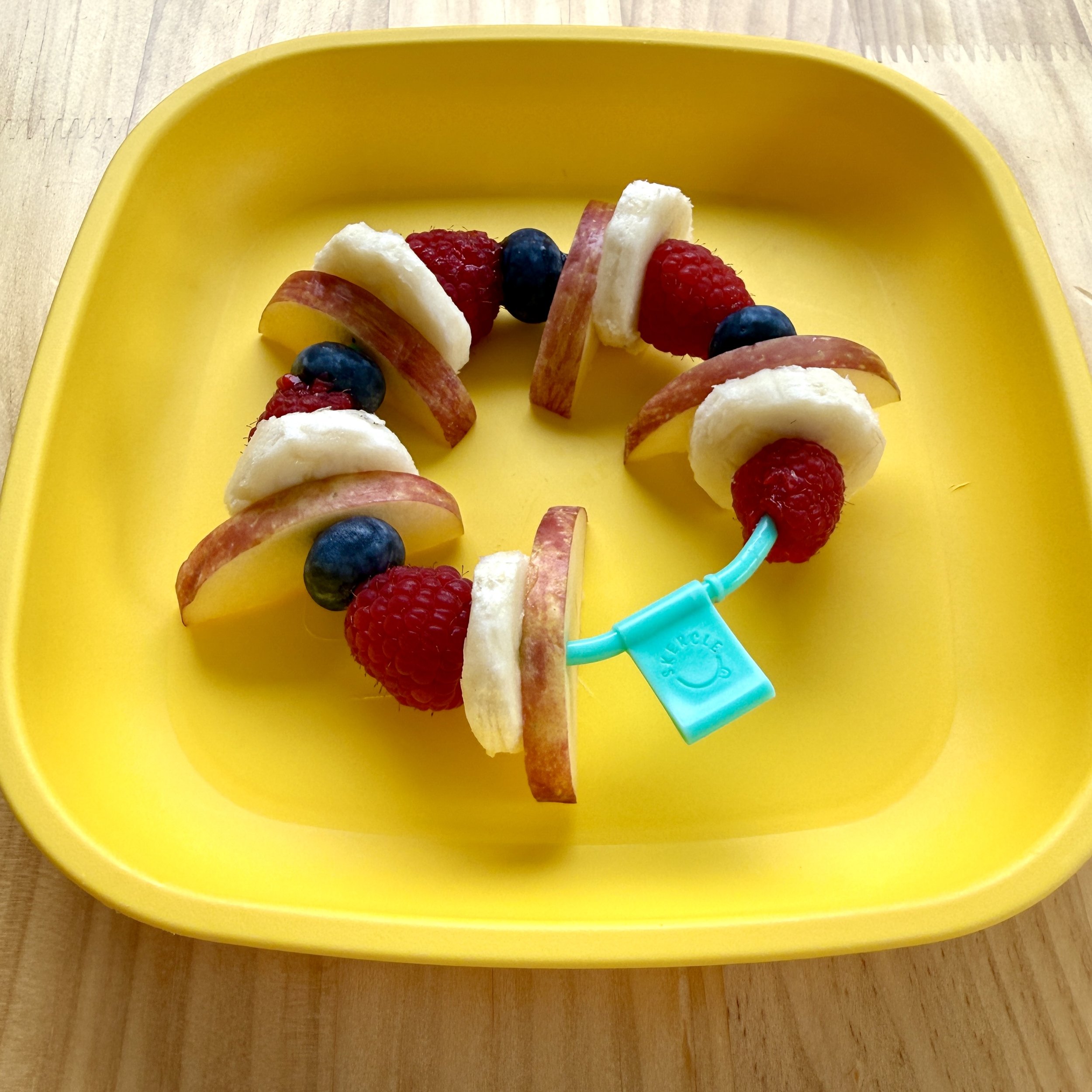 A yellow bowl containing a fruit skewer with slices of apple, banana, raspberry, and blueberry, placed on a wooden surface.