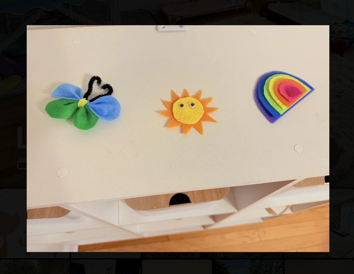 Colorful felt craft items including a butterfly with black pipe cleaner antennae and green, blue, and purple wings, a smiling sun with orange rays and a yellow face with googly eyes, and a rainbow with rainbow-colored felt layers.