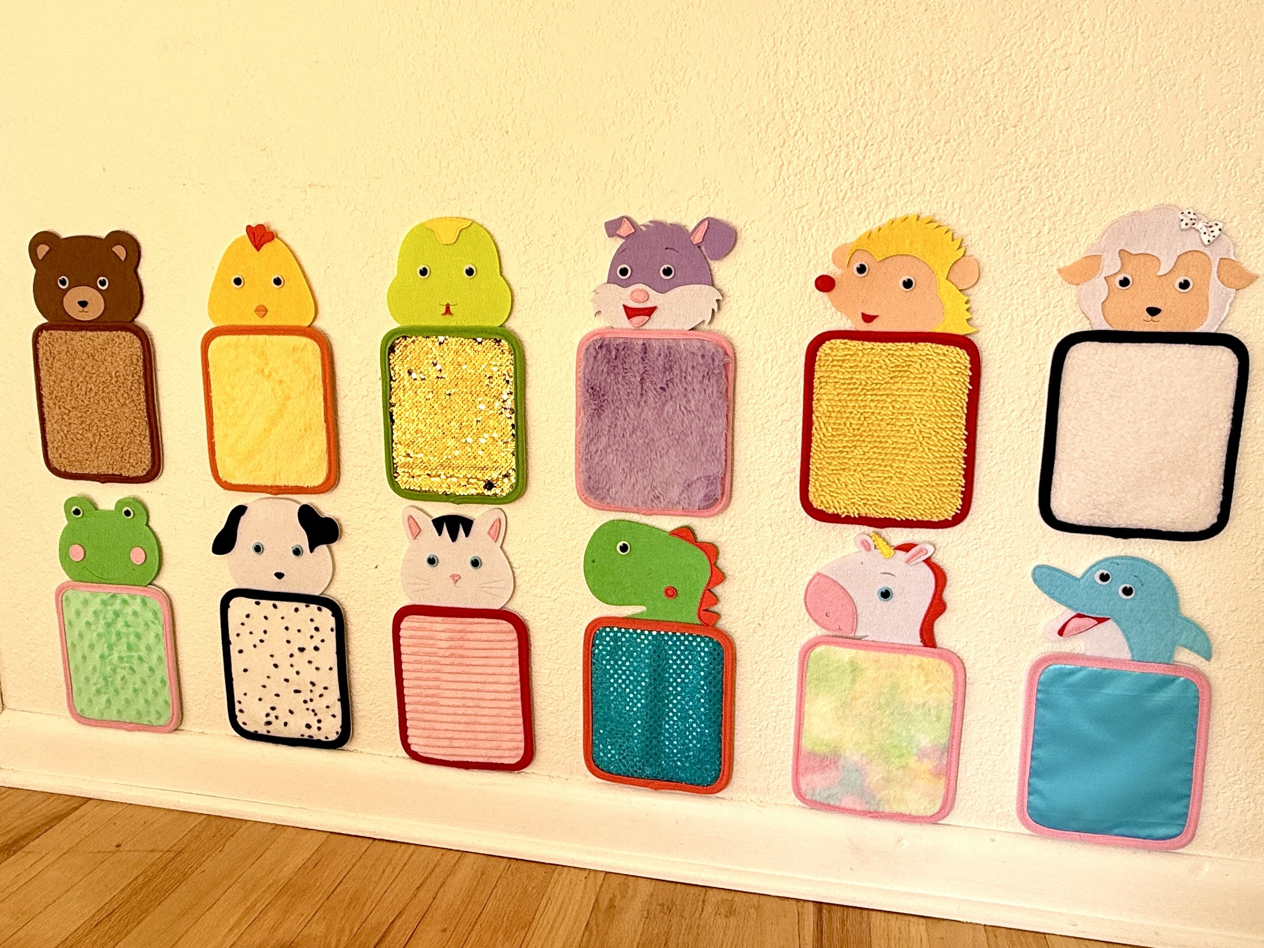 Colorful felt sensory display with various animal-shaped felt pieces and rectangular textured mats, arranged on a cream-colored wall.