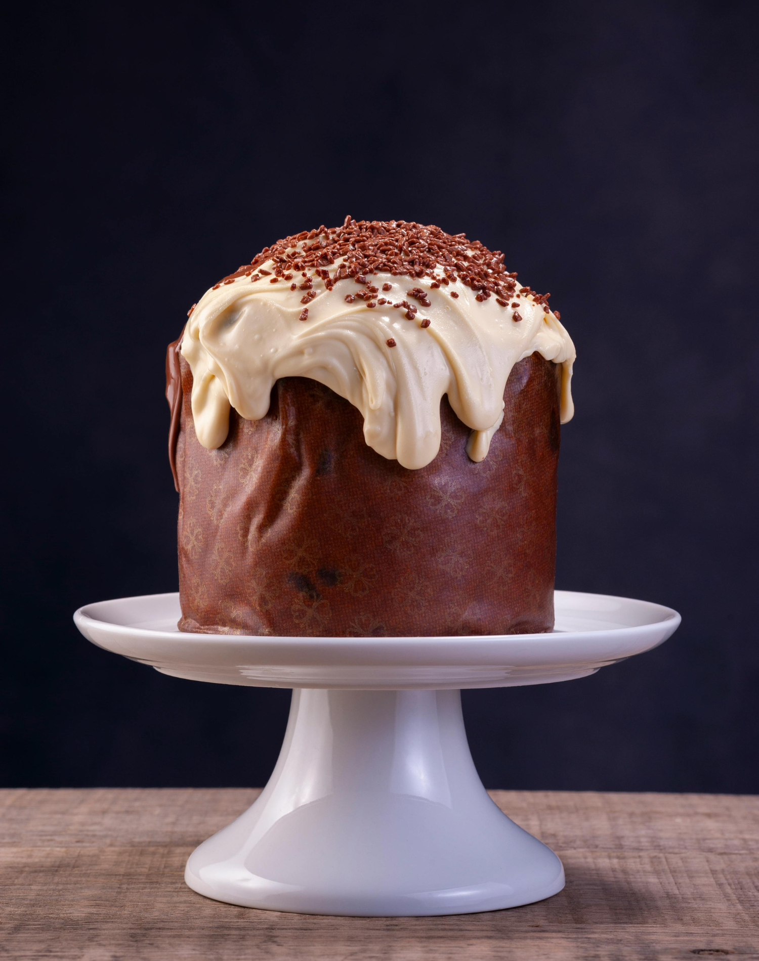 Chocolate panettone topped with white chocolate icing and sprinkles, placed on a white cake stand against a dark background.