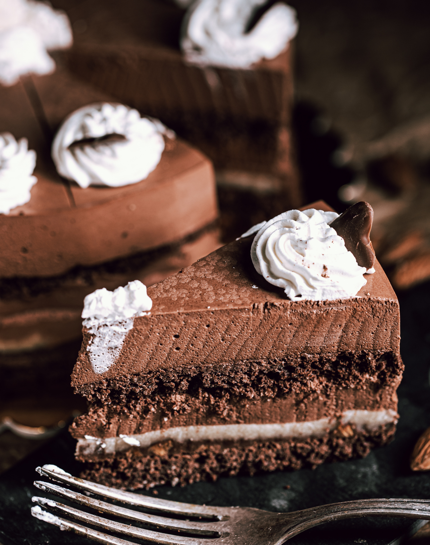A slice of chocolate cake topped with whipped cream and a small piece of chocolate on a dark plate with a fork in the foreground, and a whole chocolate cake in the background.