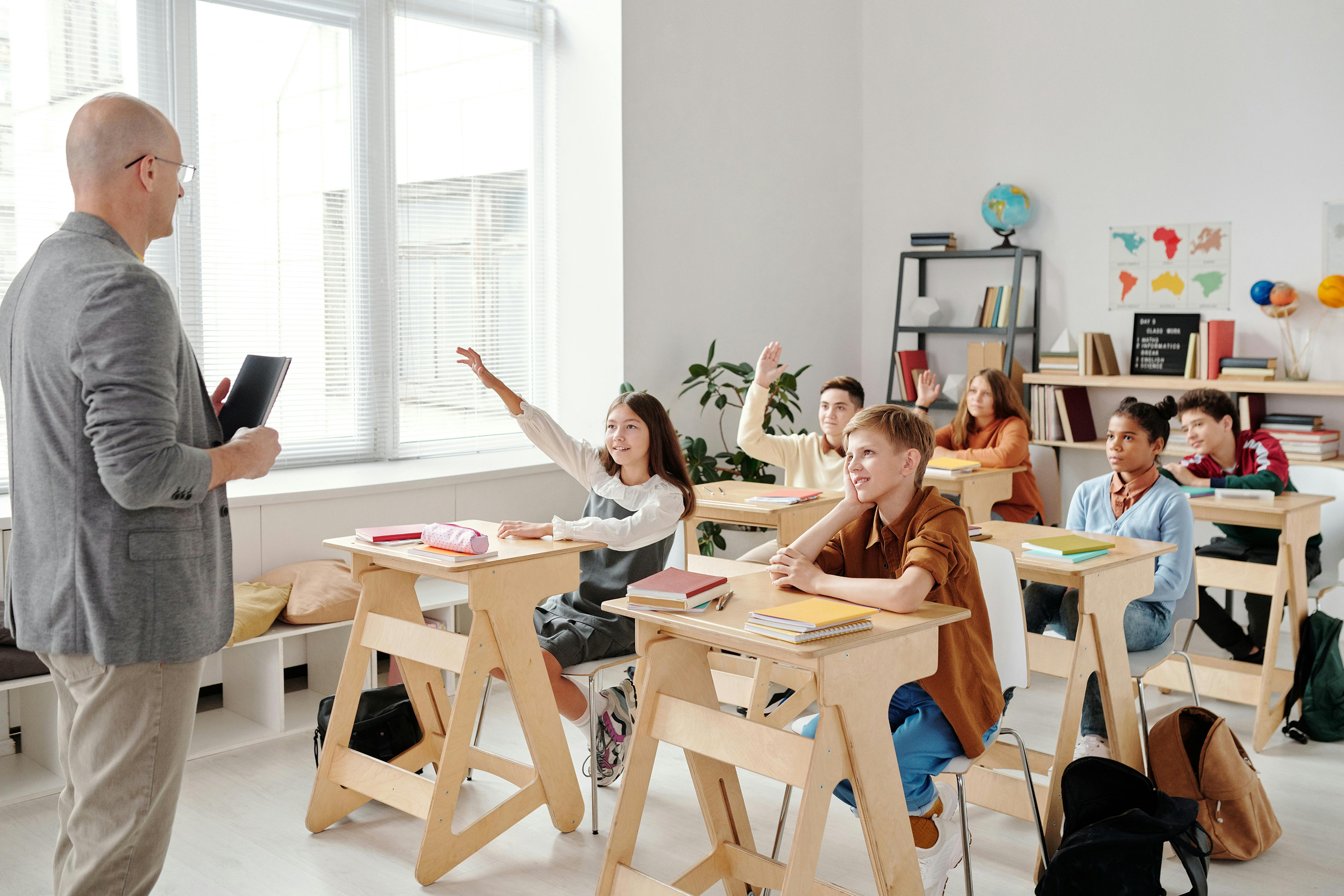 A classroom with six students raising their hands, paying attention to a teacher who is holding a clipboard, with desks, books, and educational decorations.