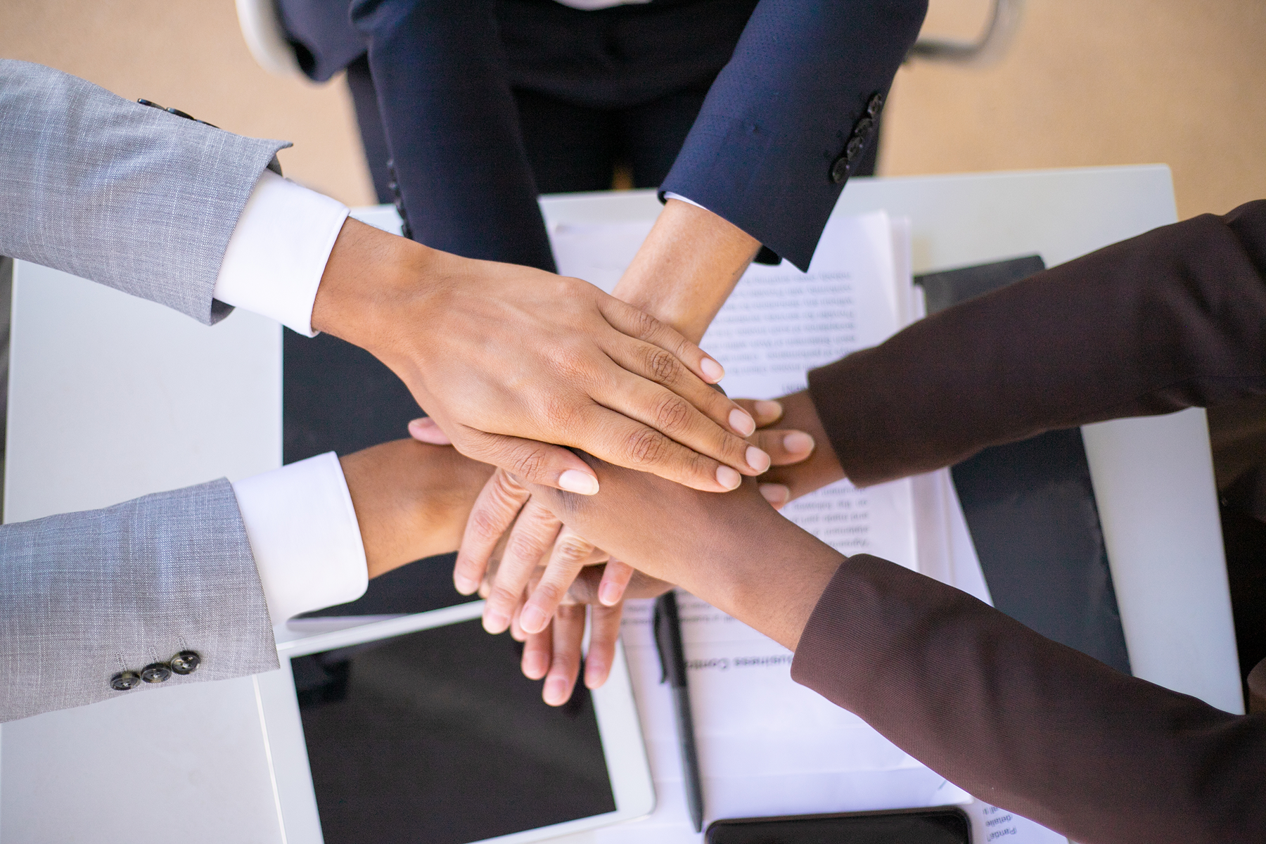 Four diverse professionals place their hands on top of each other in a show of teamwork, above a desk with a tablet, pen, and documents.