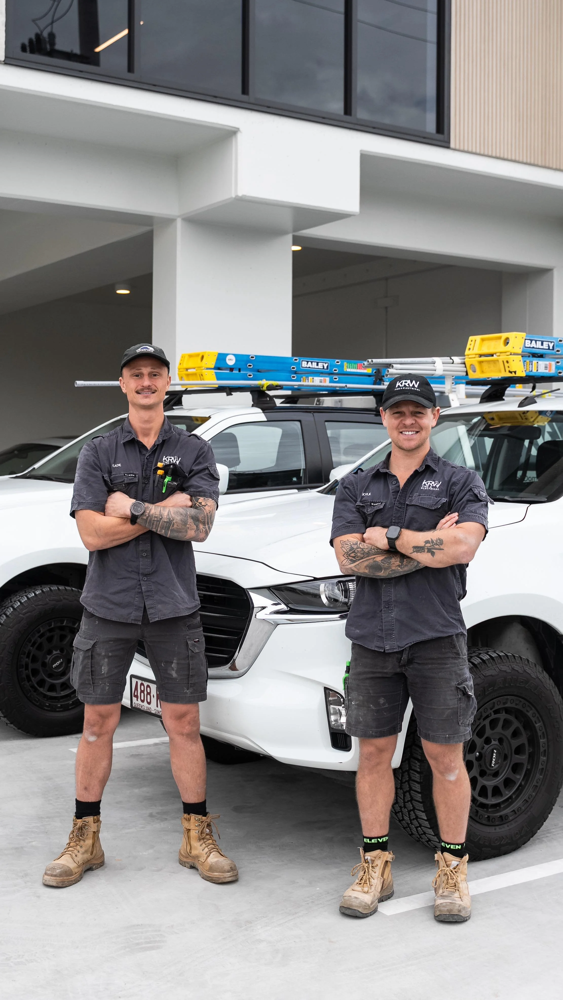Two men in work uniforms standing with arms crossed in front of a white service vehicle, with ladders on top, in a parking lot outside a modern building.
