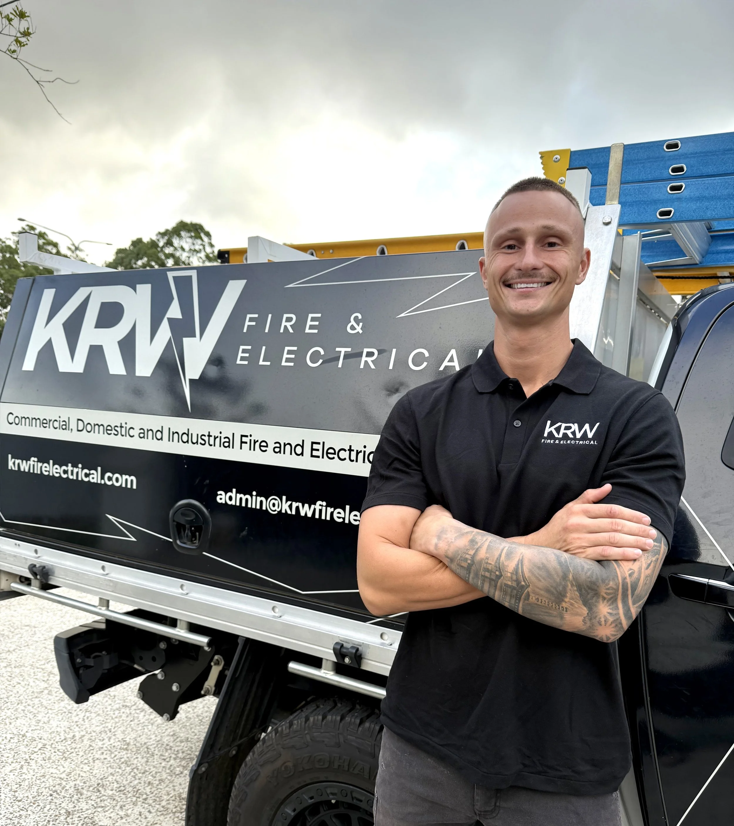 A man standing in front of a Krw Fire & Electrical truck, smiling with arms crossed, wearing a black polo shirt with the company's logo.