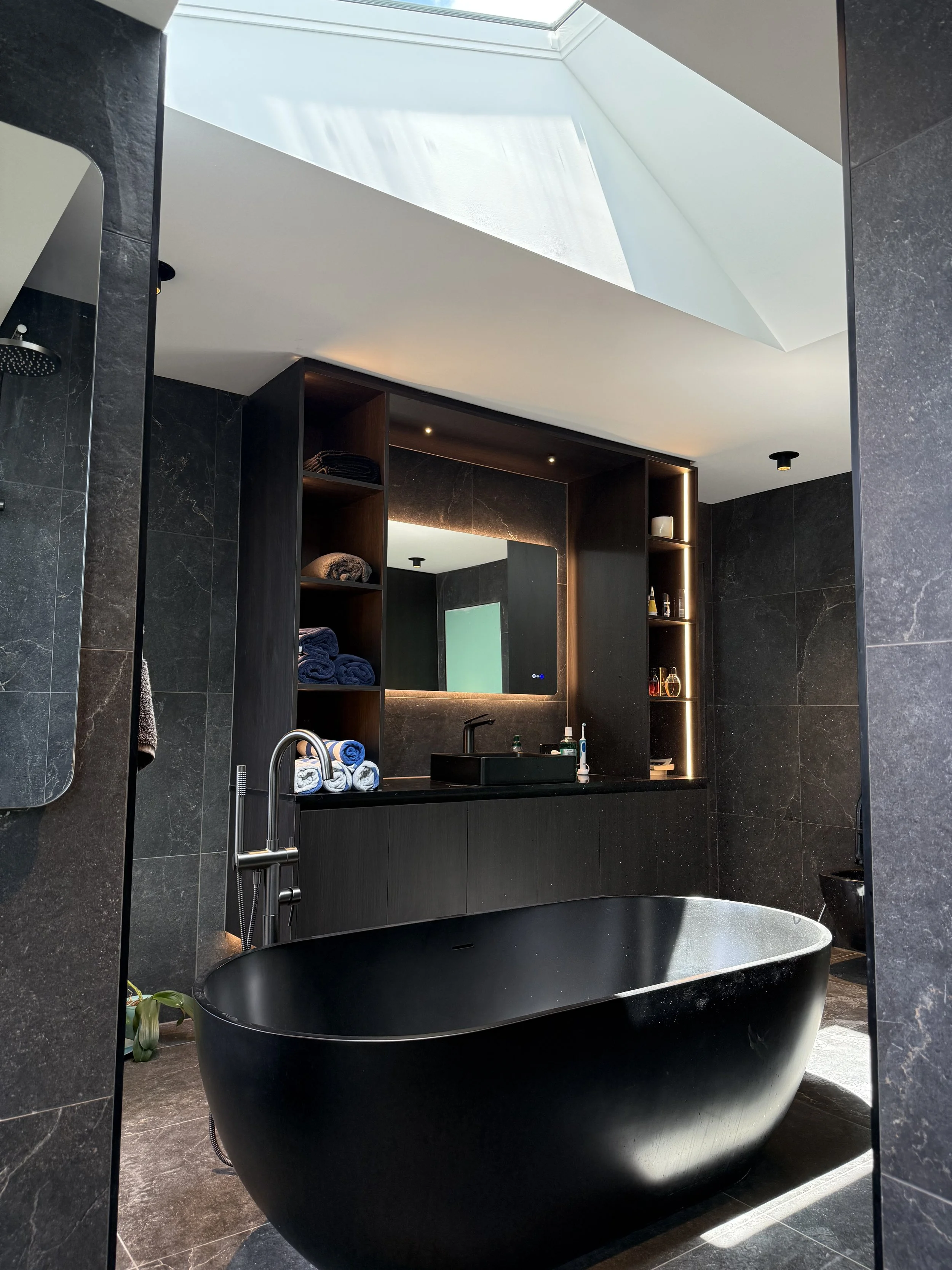 Modern bathroom with black bathtub, dark wood vanity, illuminated shelves, and a large skylight.