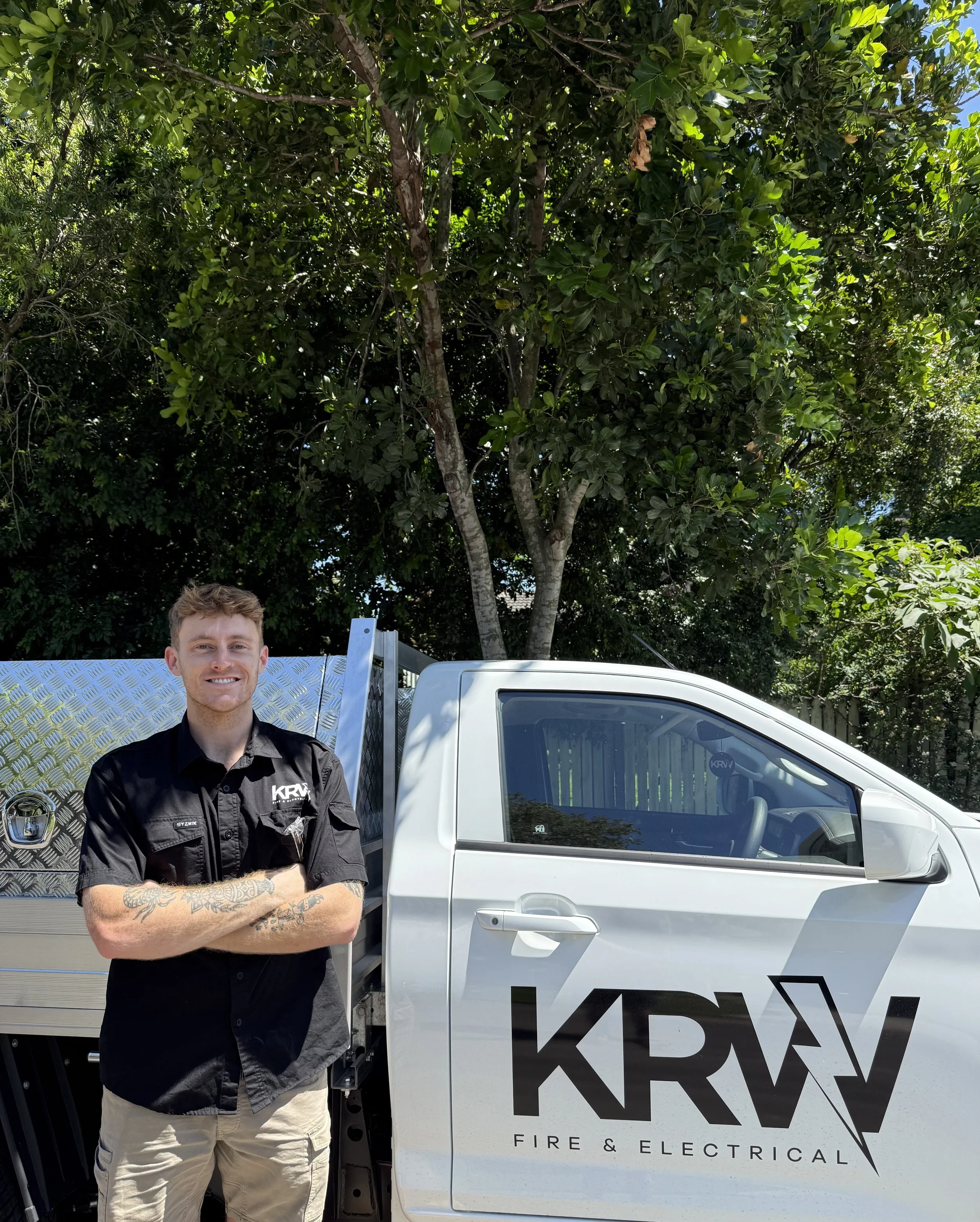 A smiling man with tattoos on his arms, wearing a black shirt with the logo 'KRAV FIRE & ELECTRICAL', standing with arms crossed next to a white utility truck with the logo 'KRAV FIRE & ELECTRICAL'. The background features a large green tree under a clear sky.
