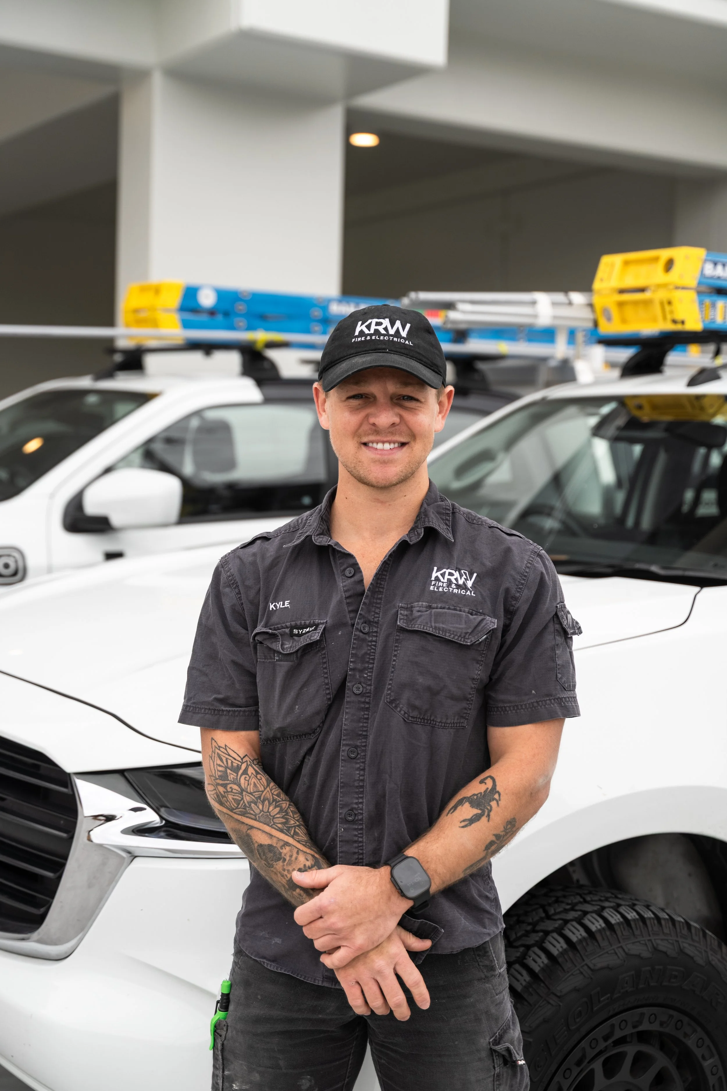 A man wearing a black KRW Fire & Electrical uniform and hat, standing in front of white service vehicles with ladders on top, smiling at the camera.
