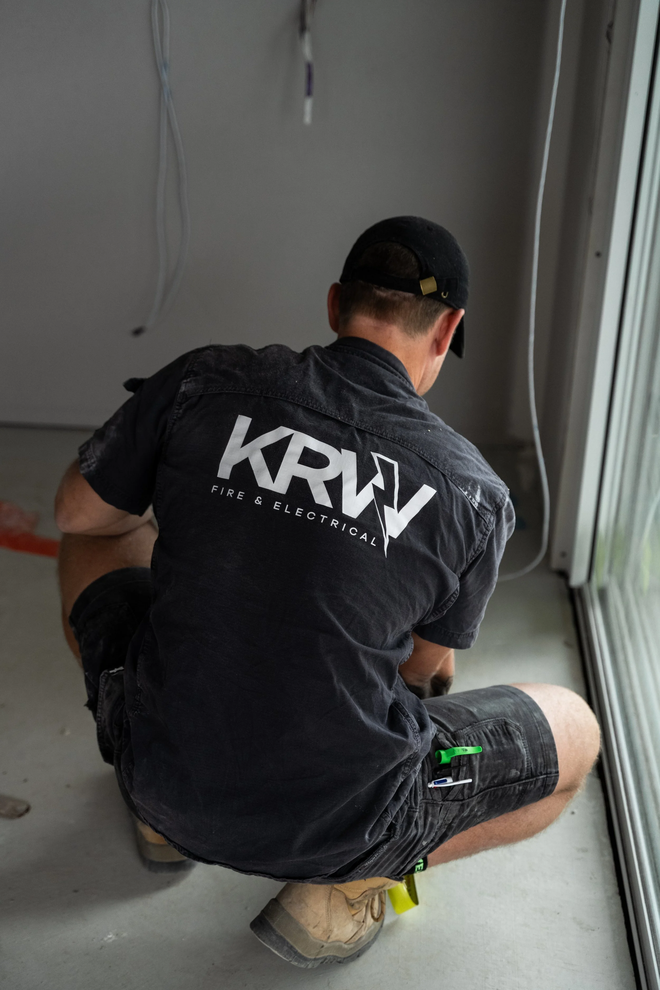 A man wearing a black cap and a black shirt with 'KRW Fire & Electrical' on the back, kneeling on the floor working on electrical wiring near a sliding glass door.
