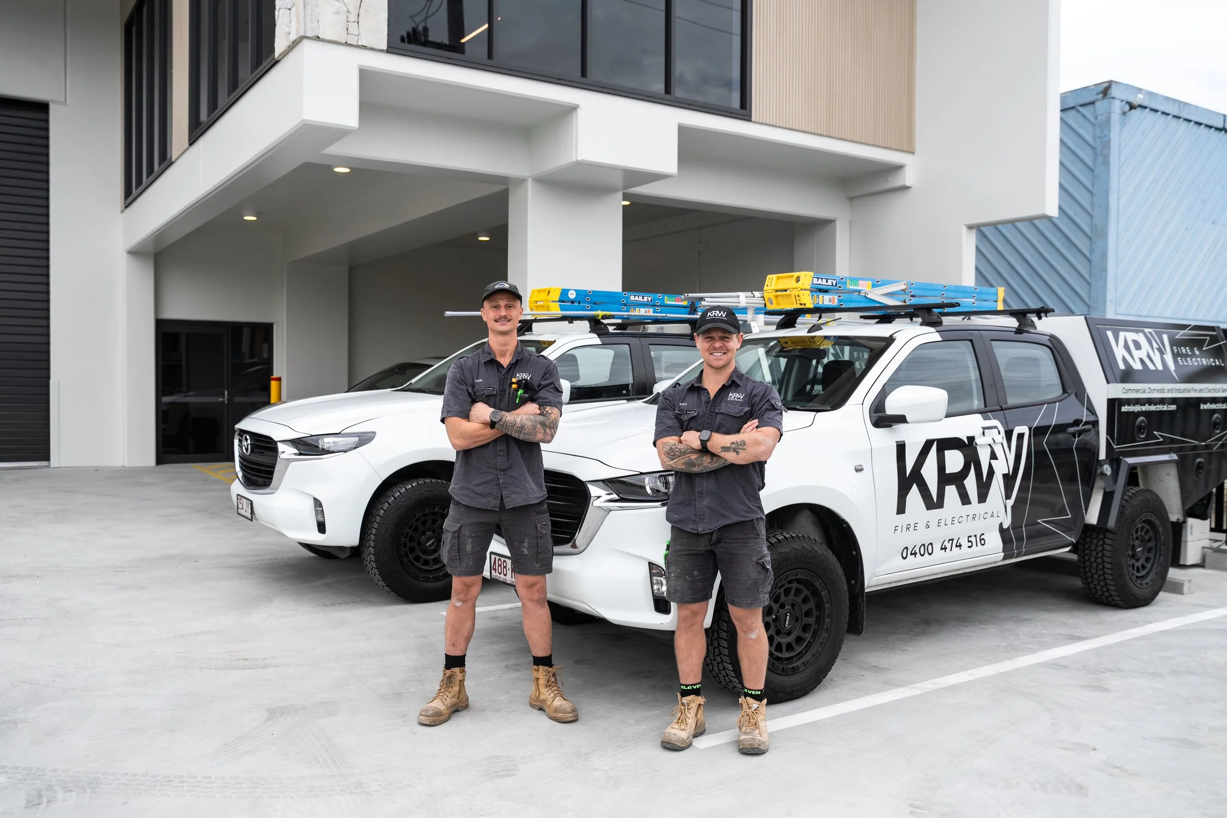 Two men in work uniforms standing with arms crossed in front of company vehicles, one white and one black, parked outside a modern building. The vehicles are labeled "KRW Fire & Electrical" and have ladders mounted on top.