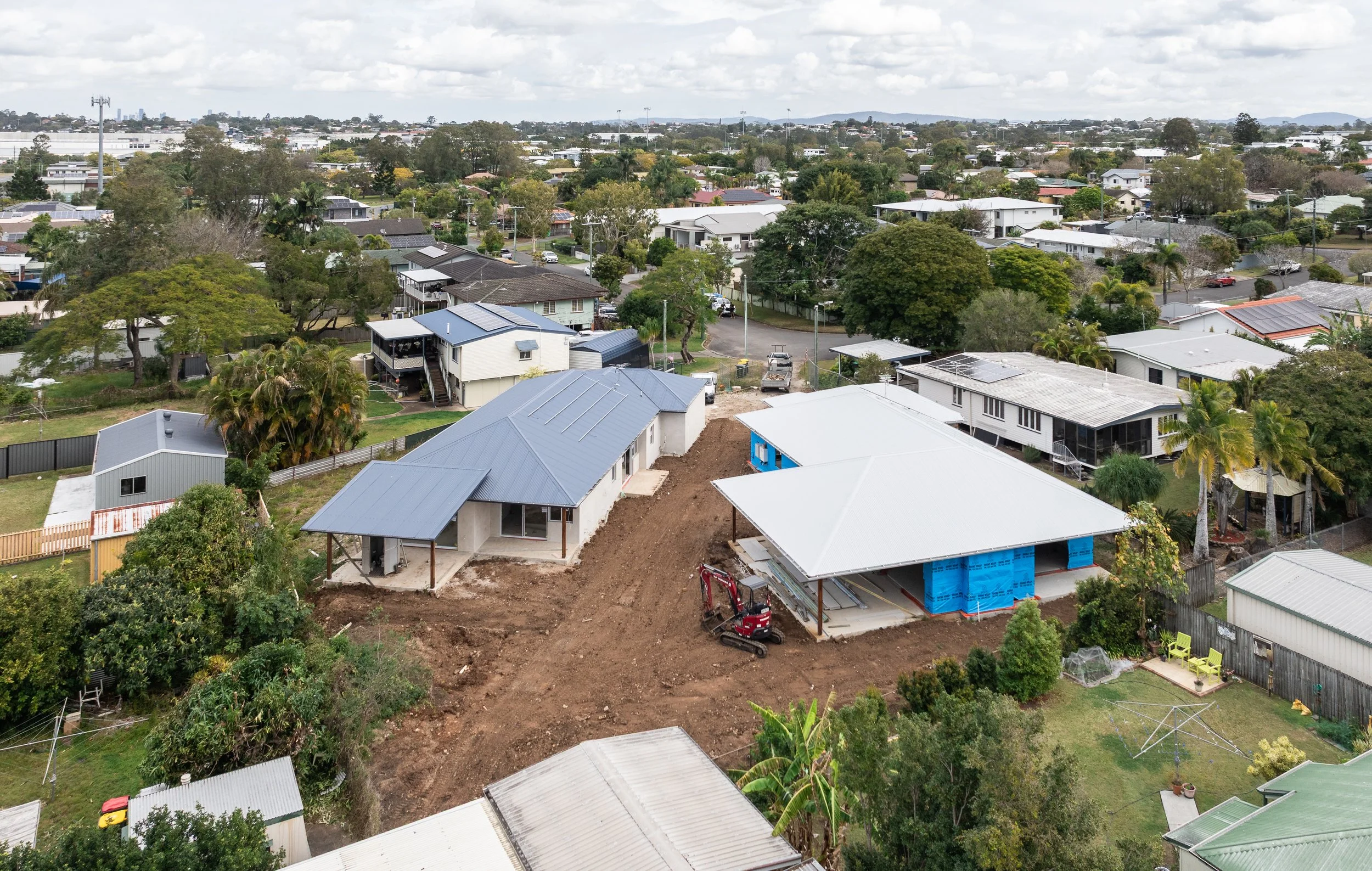 Aerial view of a residential neighborhood with houses, trees, and streets, featuring construction work on new houses with dirt and a small excavator.