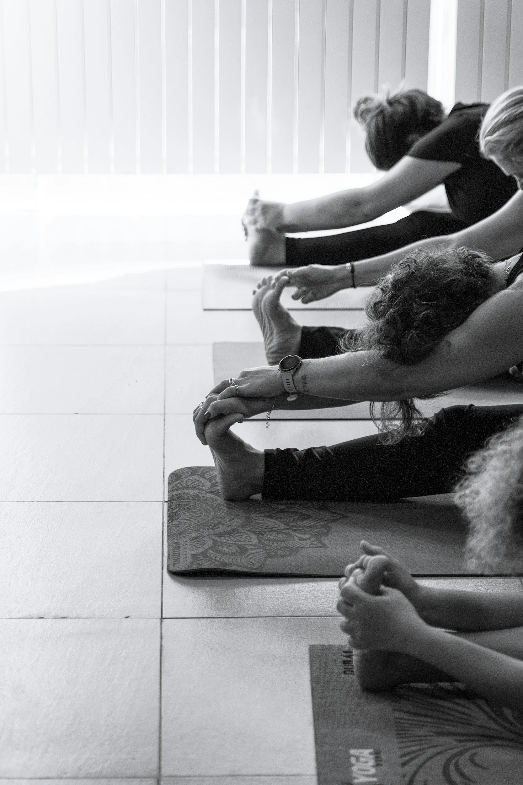 A group of people practicing yoga indoors, sitting on yoga mats with their legs extended and holding their feet with both hands, near a window with vertical blinds.