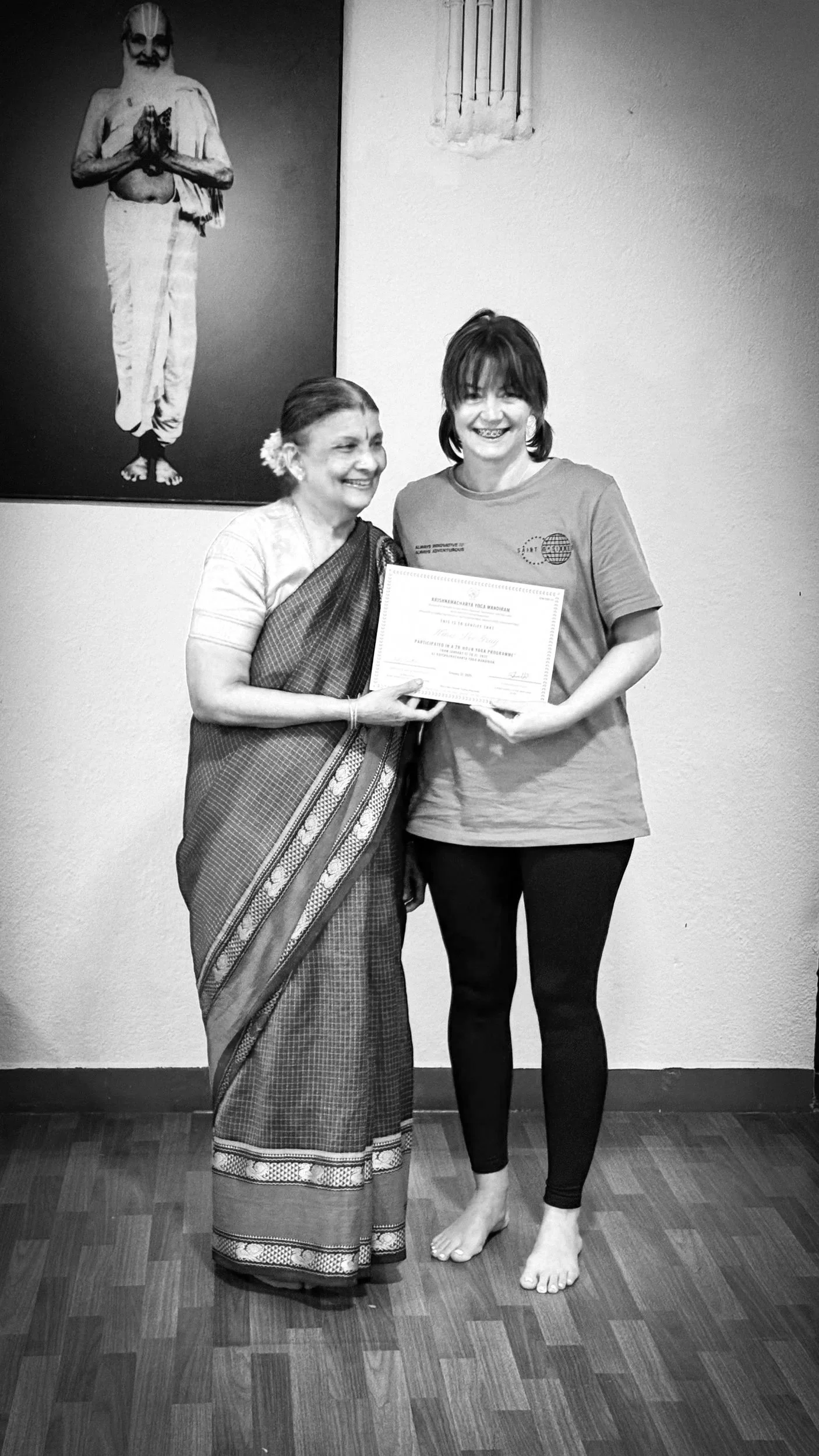 An elderly woman dressed in traditional Indian attire and a younger woman in a t-shirt and leggings are smiling while holding a certificate together. A large picture of a man in traditional Indian attire is hanging on the wall behind them.